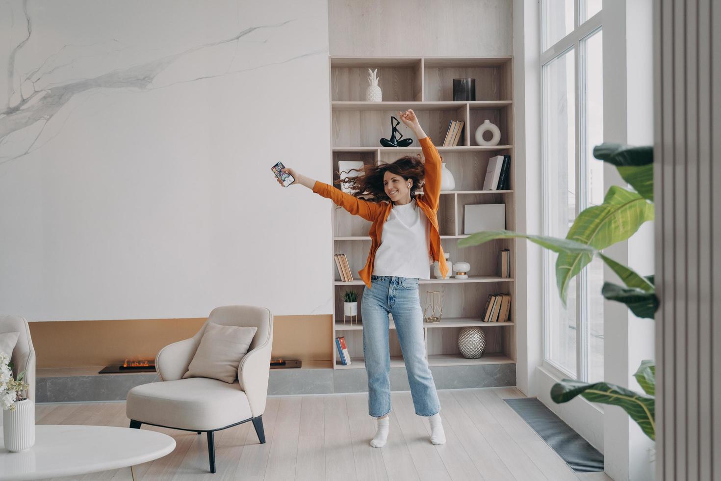 mujer enérgica bailando en la sala de estar con música de un smartphone en el moderno interior de un apartamento foto