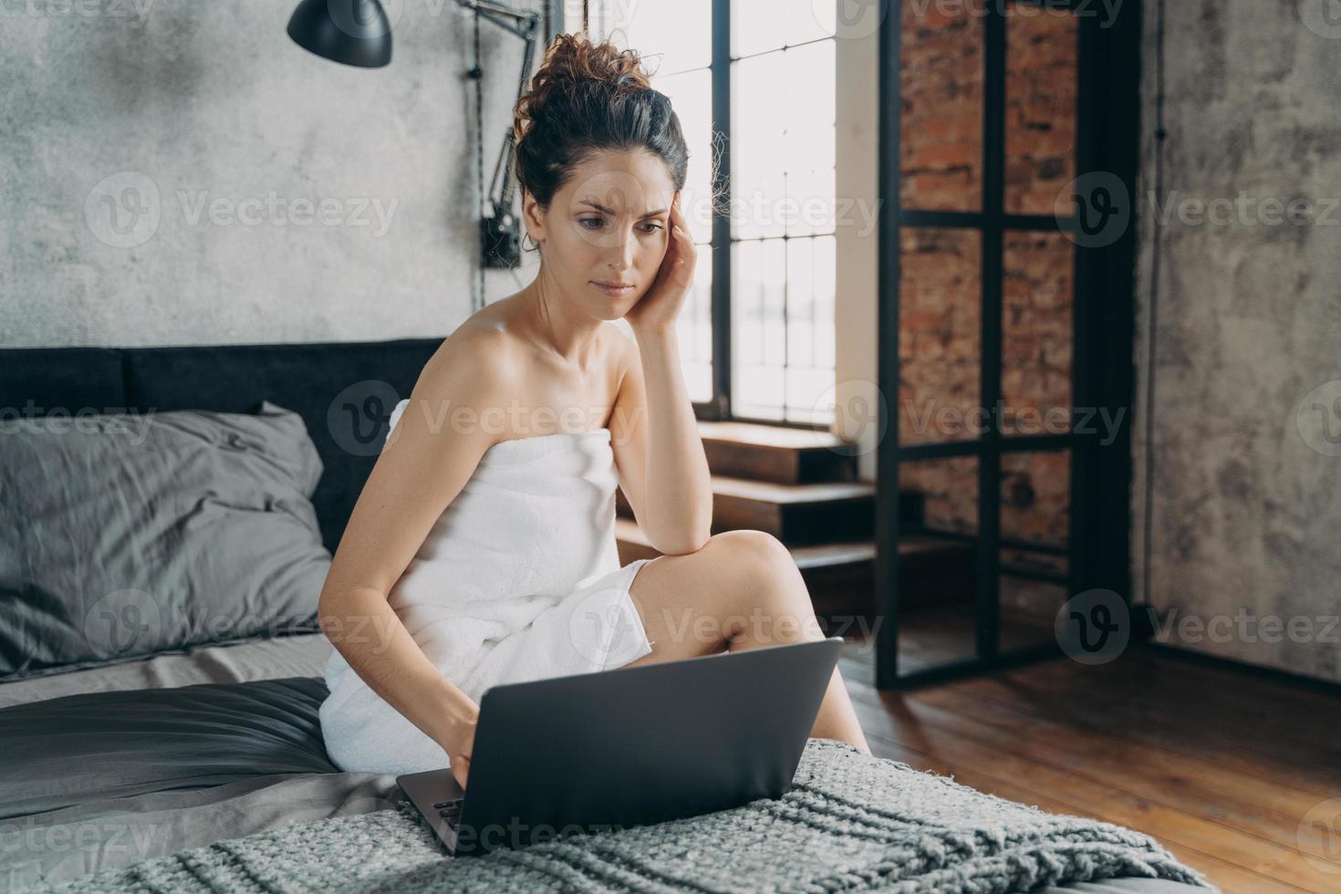 una mujer de negocios enfocada trabaja en una laptop sentada en la cama después de la ducha por la mañana en casa. trabajo remoto foto