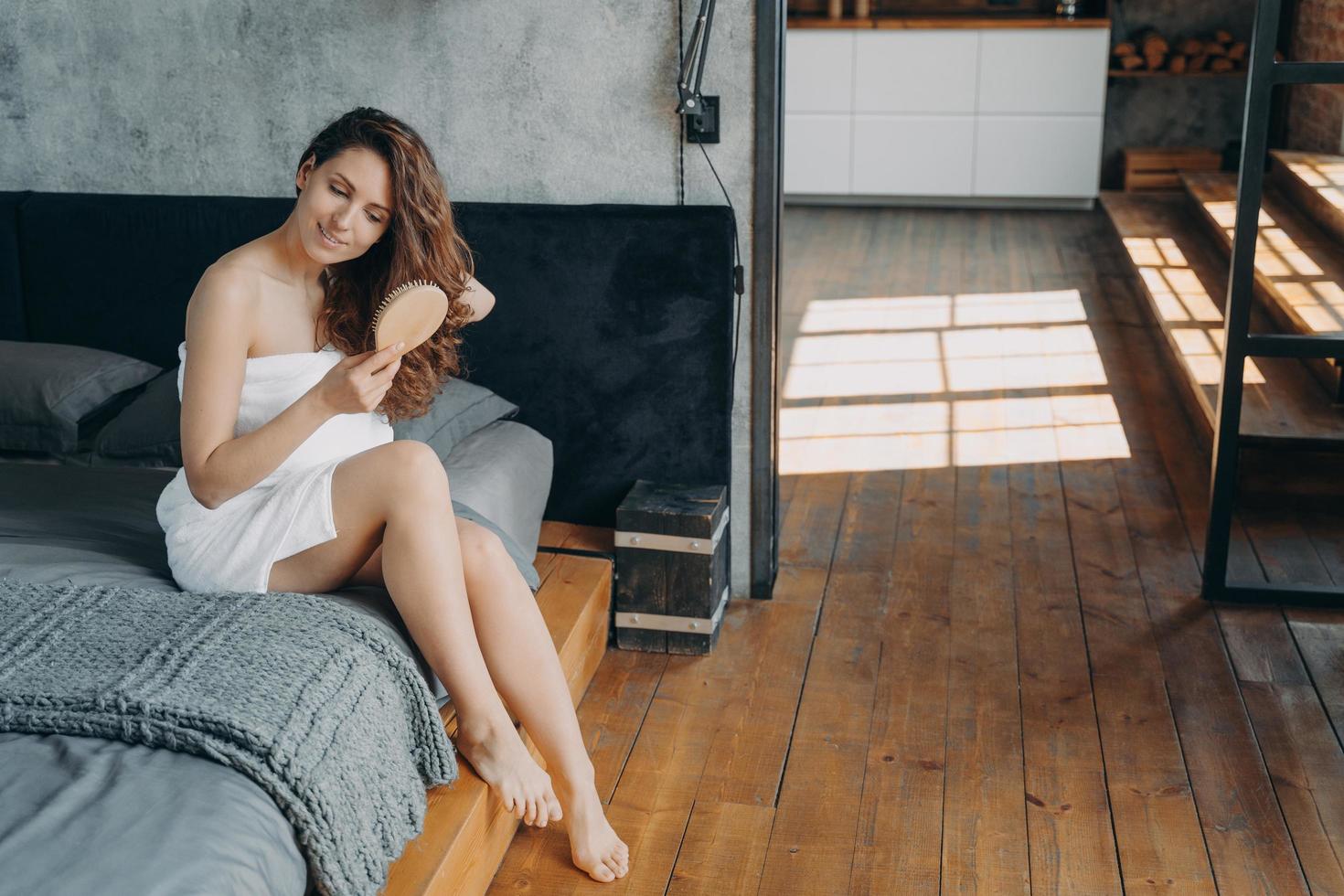 Girl wrapped in towel brushing long hair after shower in bedroom. Haircare cosmetics advertising photo