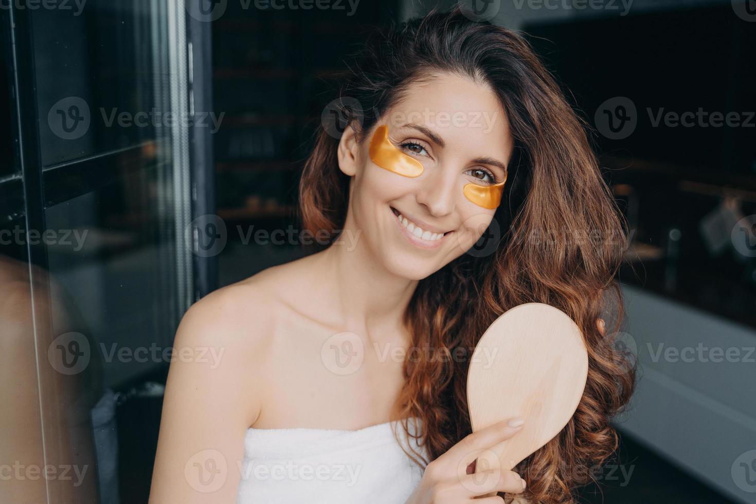 Young spanish girl with under eye patches brushes hair after shower. Morning beauty routine at home photo