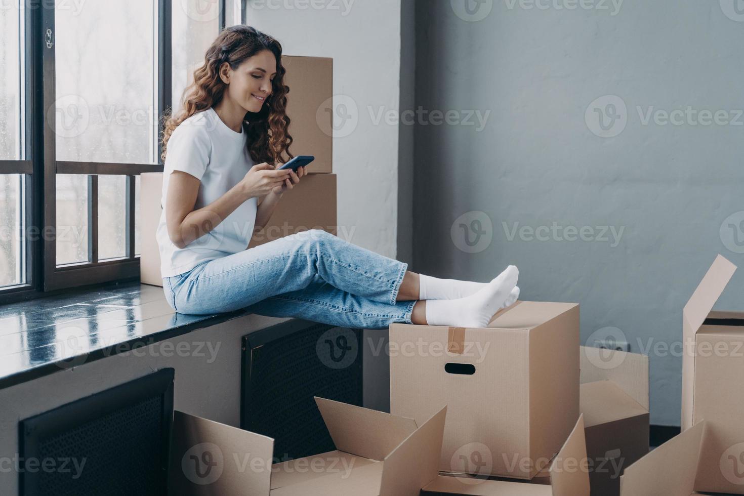 Happy girl using smartphone apps, choosing moving company, sitting with boxes. Relocation, new home photo