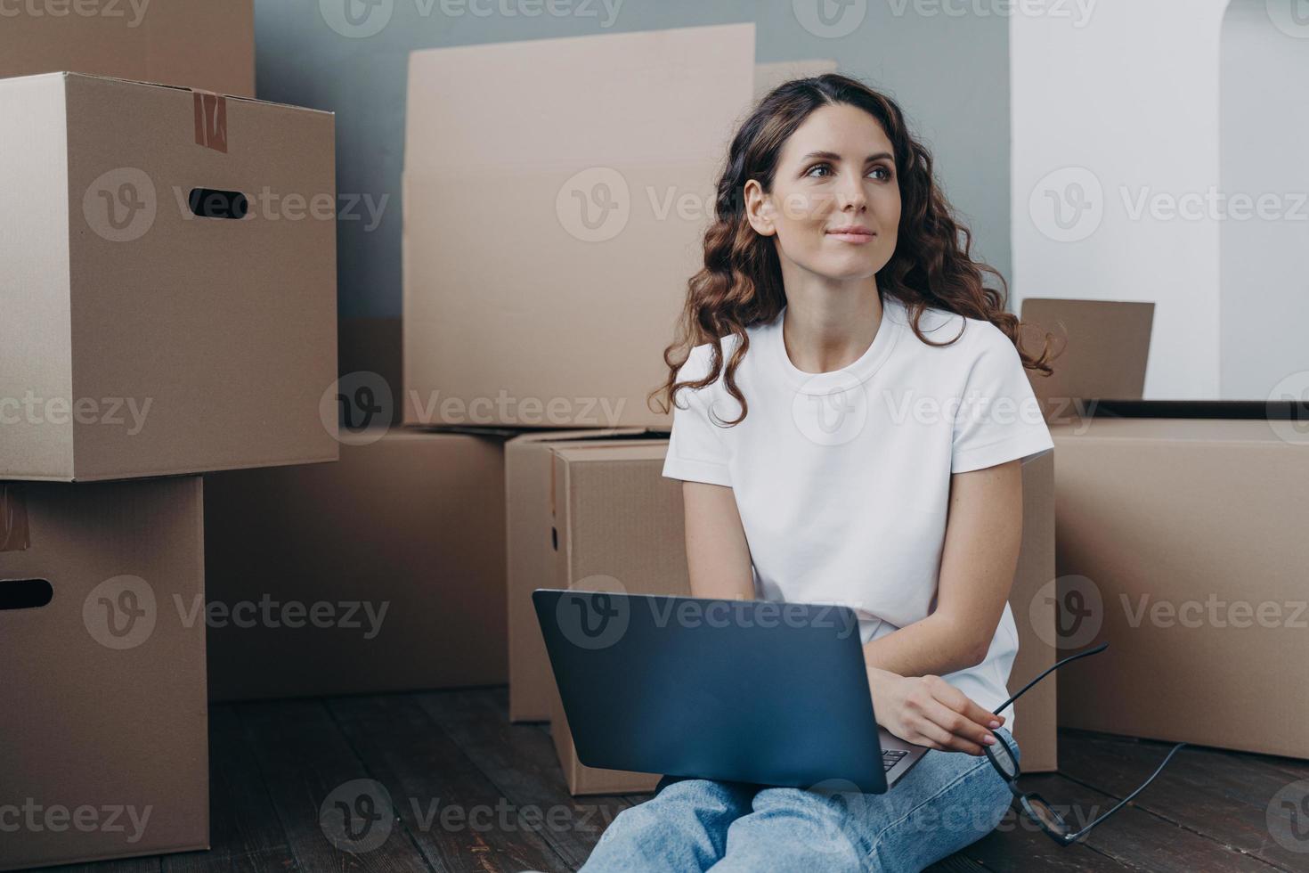 Happy girl with laptop sit on floor near cardboard boxes dreaming about new home. Relocation concept photo