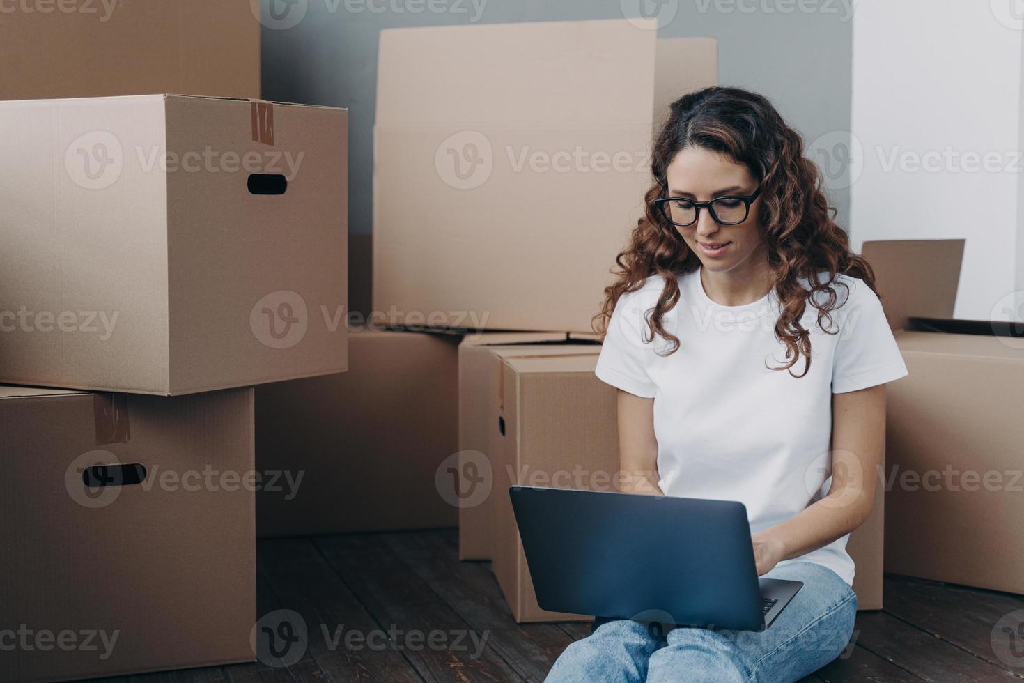 Female review moving company document on laptop online sitting on floor with boxes for relocation photo