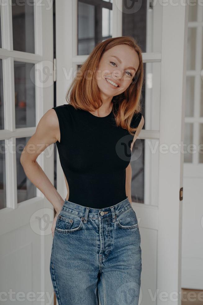Lovely redhead caucasian model in bodysuit and jeans is standing near door window and smiling. photo