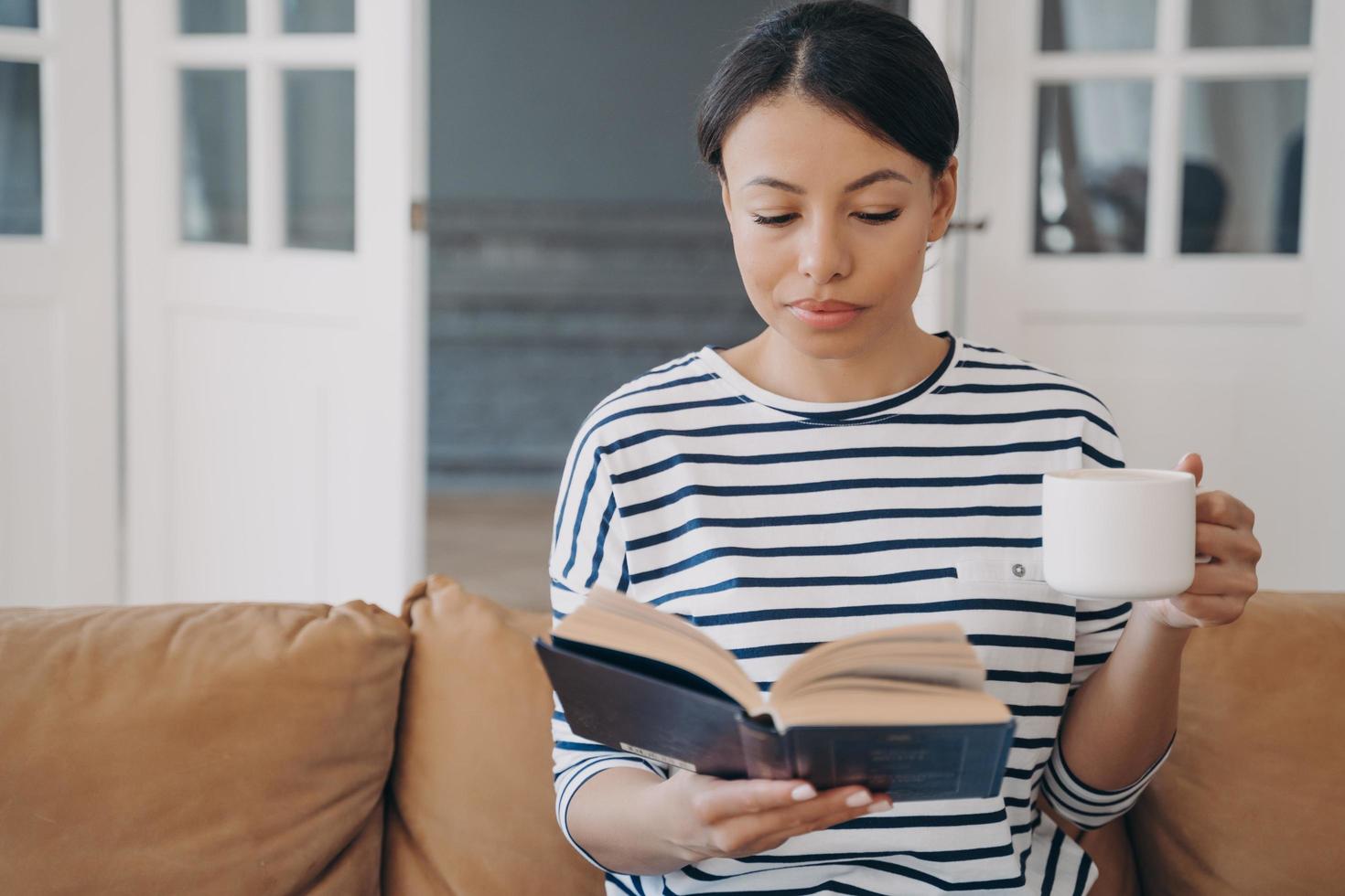 Female bookworm reading bestseller book, relaxing with coffee cup on sofa at home. Education, hobby photo