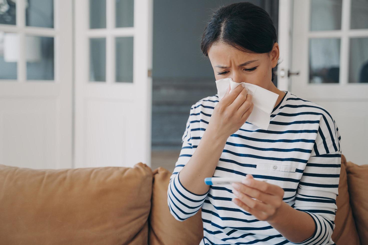 Unhealthy female blowing her nose holds thermometer, suffering from high temperature, fever at home photo