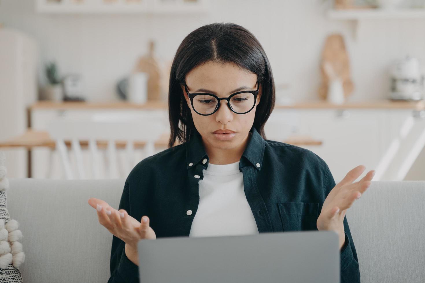 Puzzled woman freelancer in glasses spreading hands, shocked by problems in work, error on laptop photo