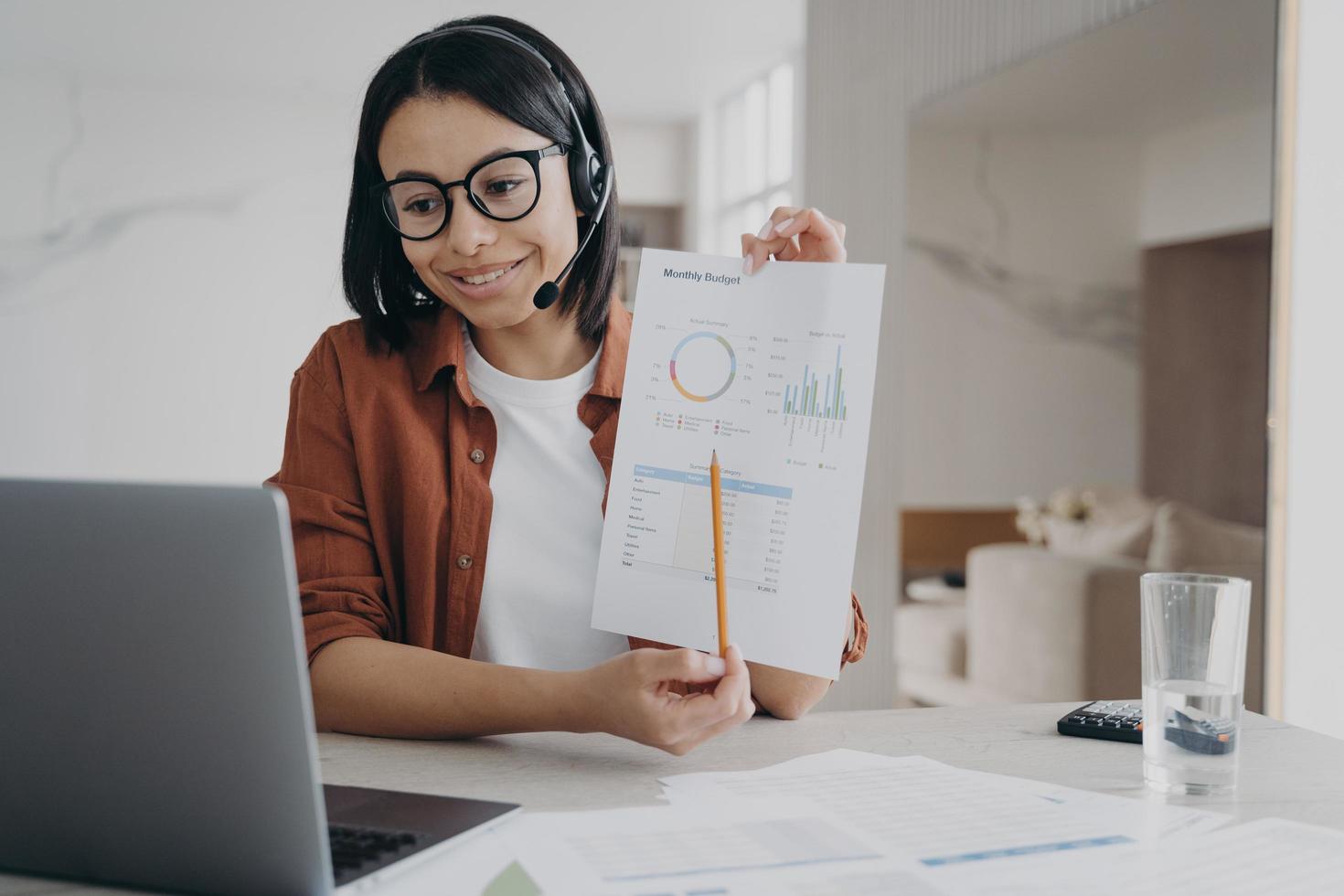 Businesswoman in headset showing chart, discussing business project while video conference at laptop photo