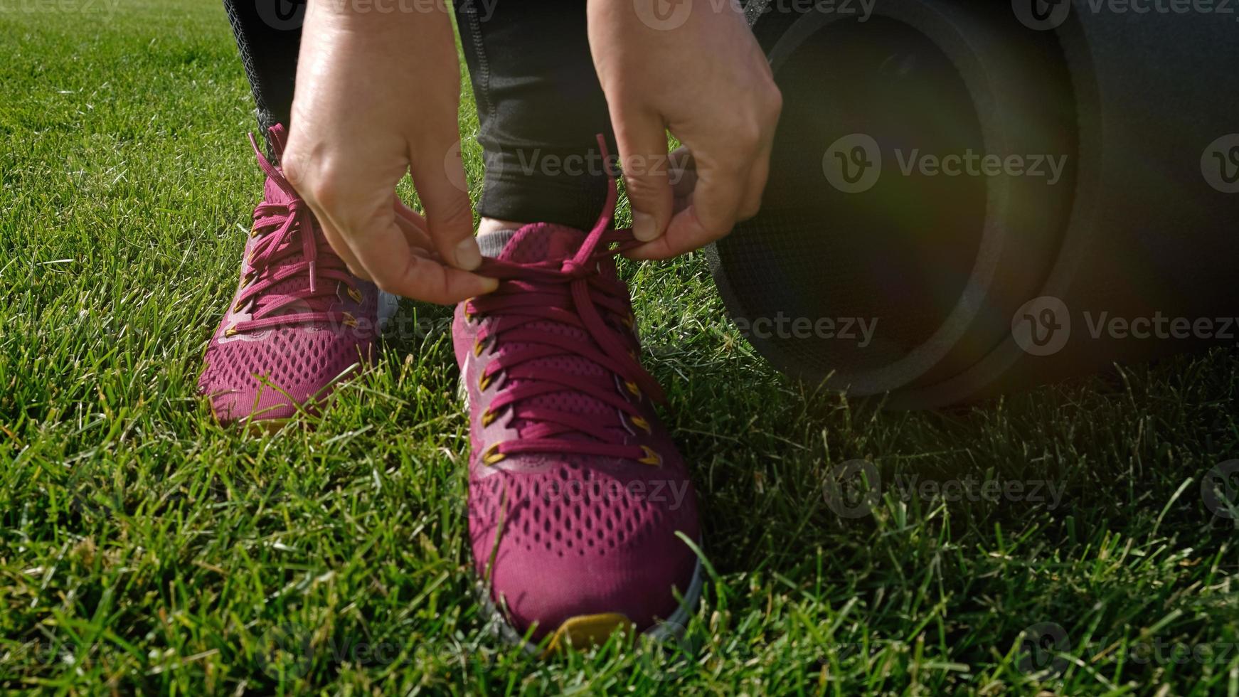 Young woman ties the laces on the pink sneaker. Woman preparing for yoga asana or fitness workout on green lawn in the backyard or park. Warm up before running. Healthy lifestyle. photo