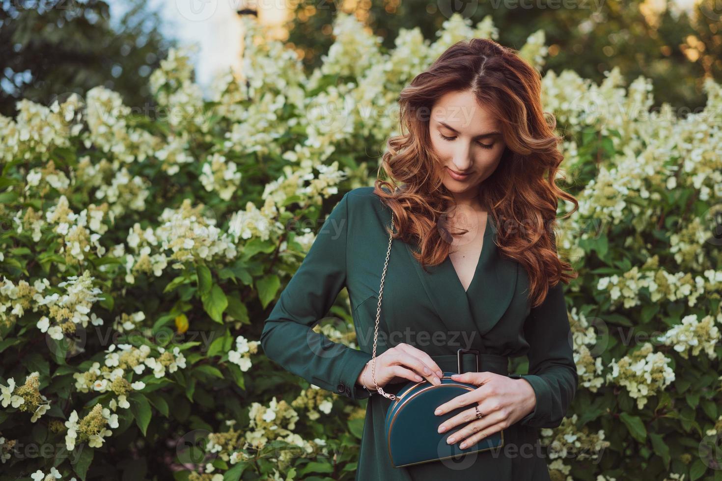 a beautiful adult woman in a green dress stands against the background of flowering bushes photo