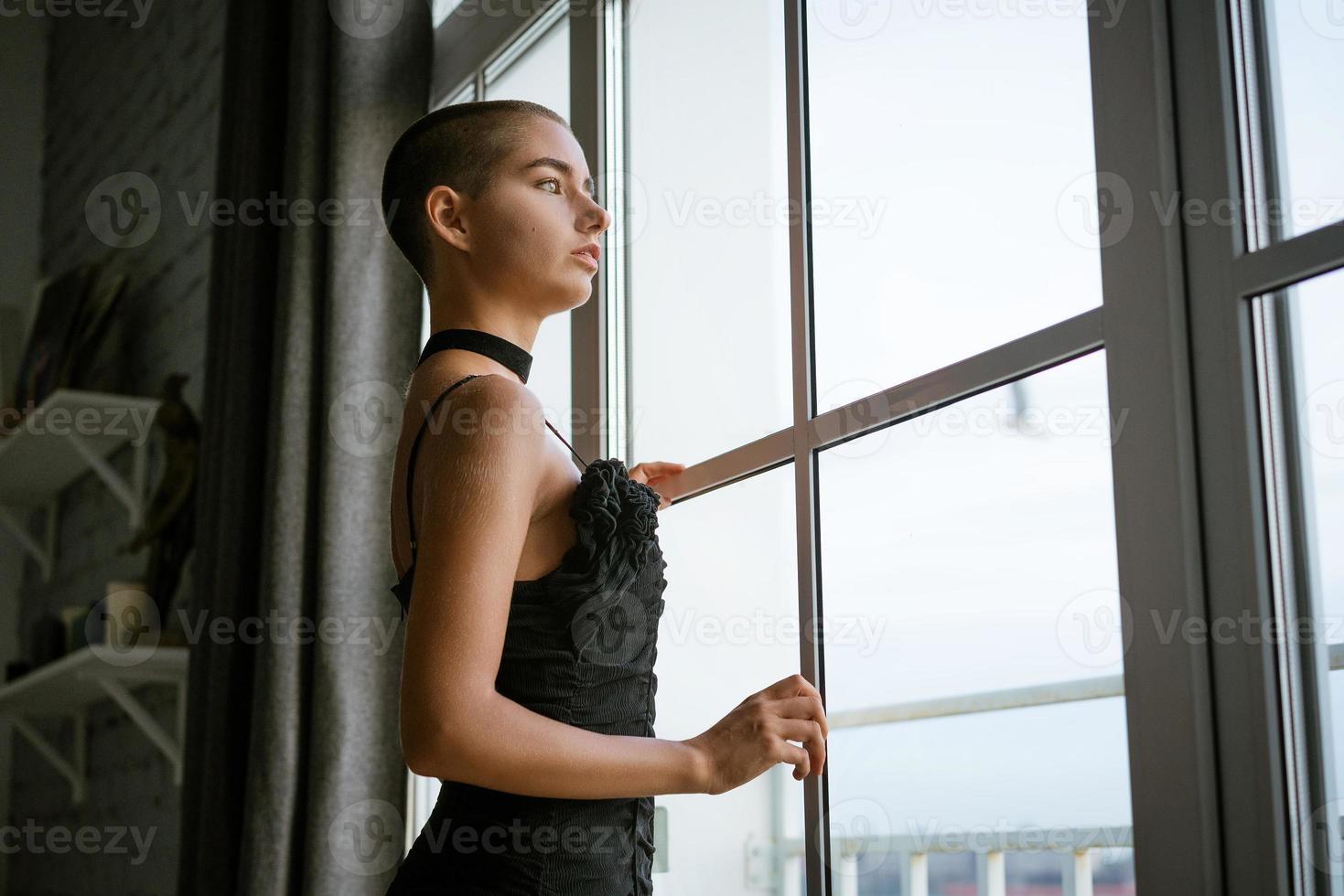 retrato de una joven triste con el pelo corto de pie mirando por la ventana con un vestido negro foto