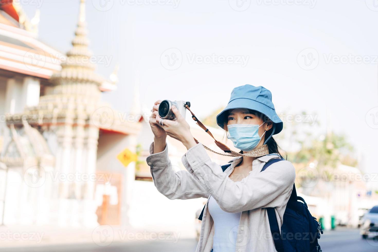 Una joven adulta asiática que viaja con mochila usa mascarilla para tomar una foto con la cámara.