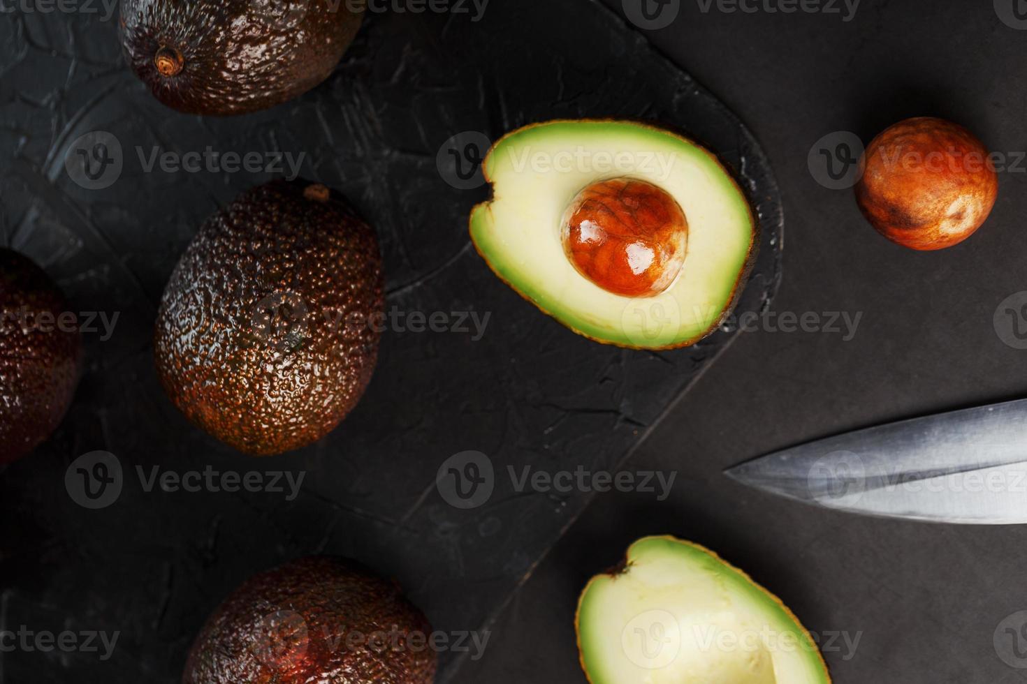 Whole and sliced avocados with a knife on a black textured black table, flat masonry. photo