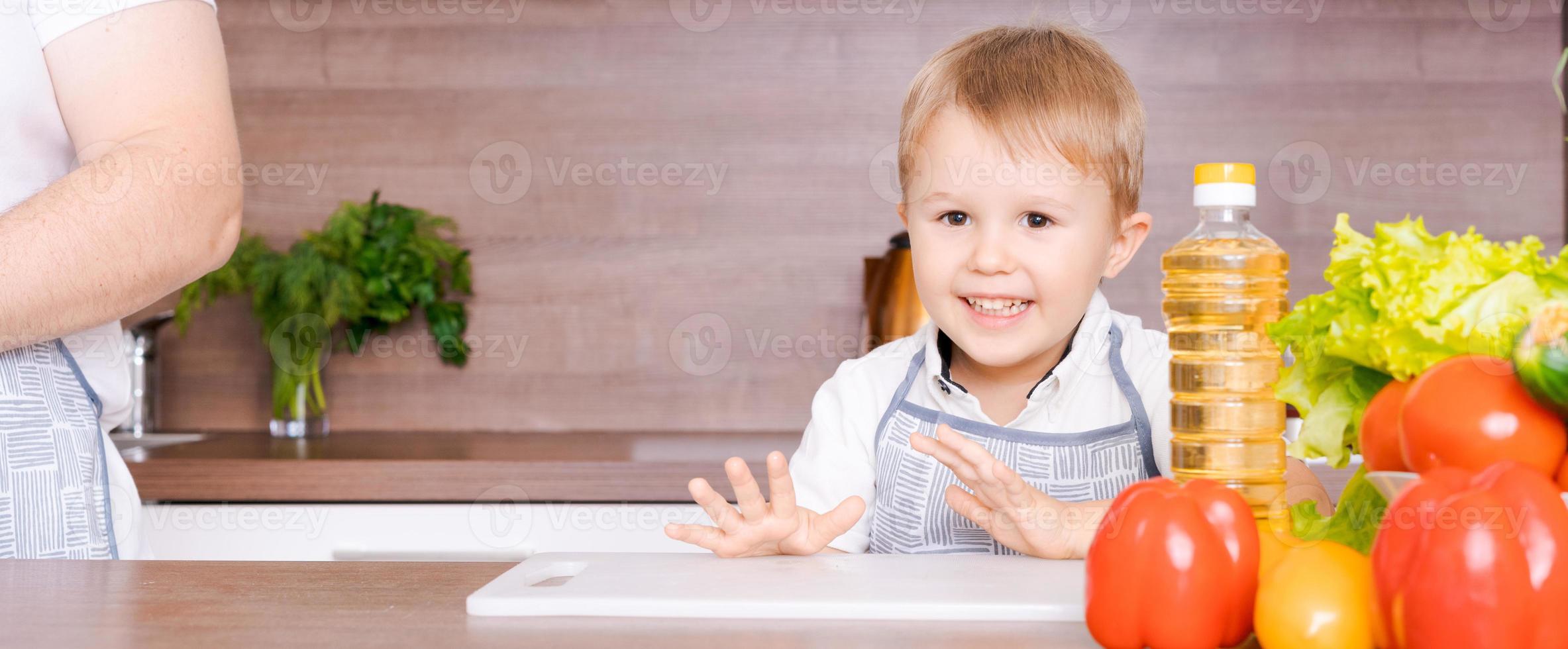 niño pequeño cocinero y un delantal en la cocina sonríe, verduras y aceite de girasol foto