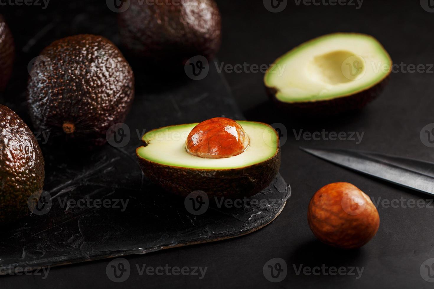 Whole and sliced avocados with a knife on a black textured black table, flat masonry. photo