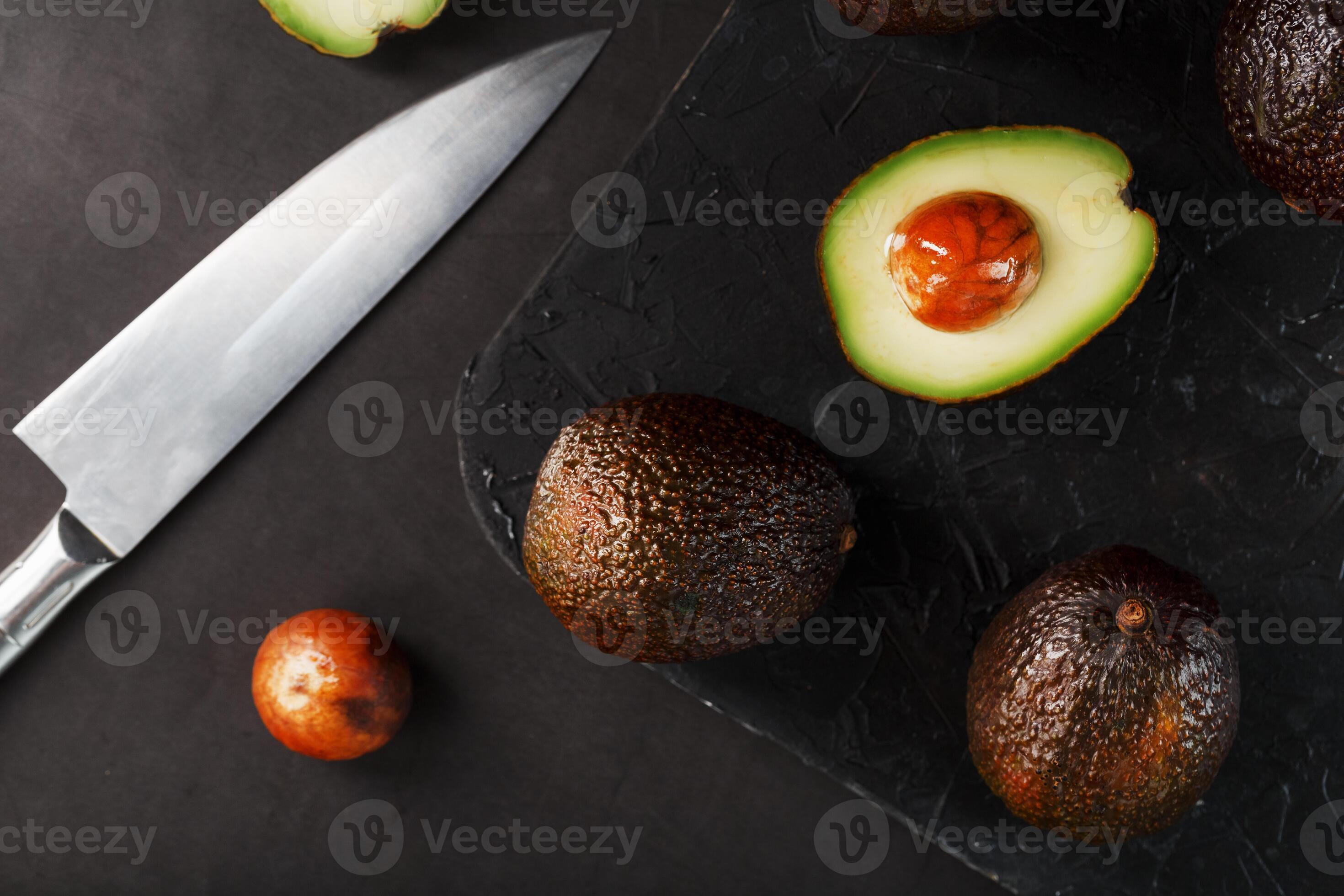 Whole and sliced avocados with a knife on a black textured black table