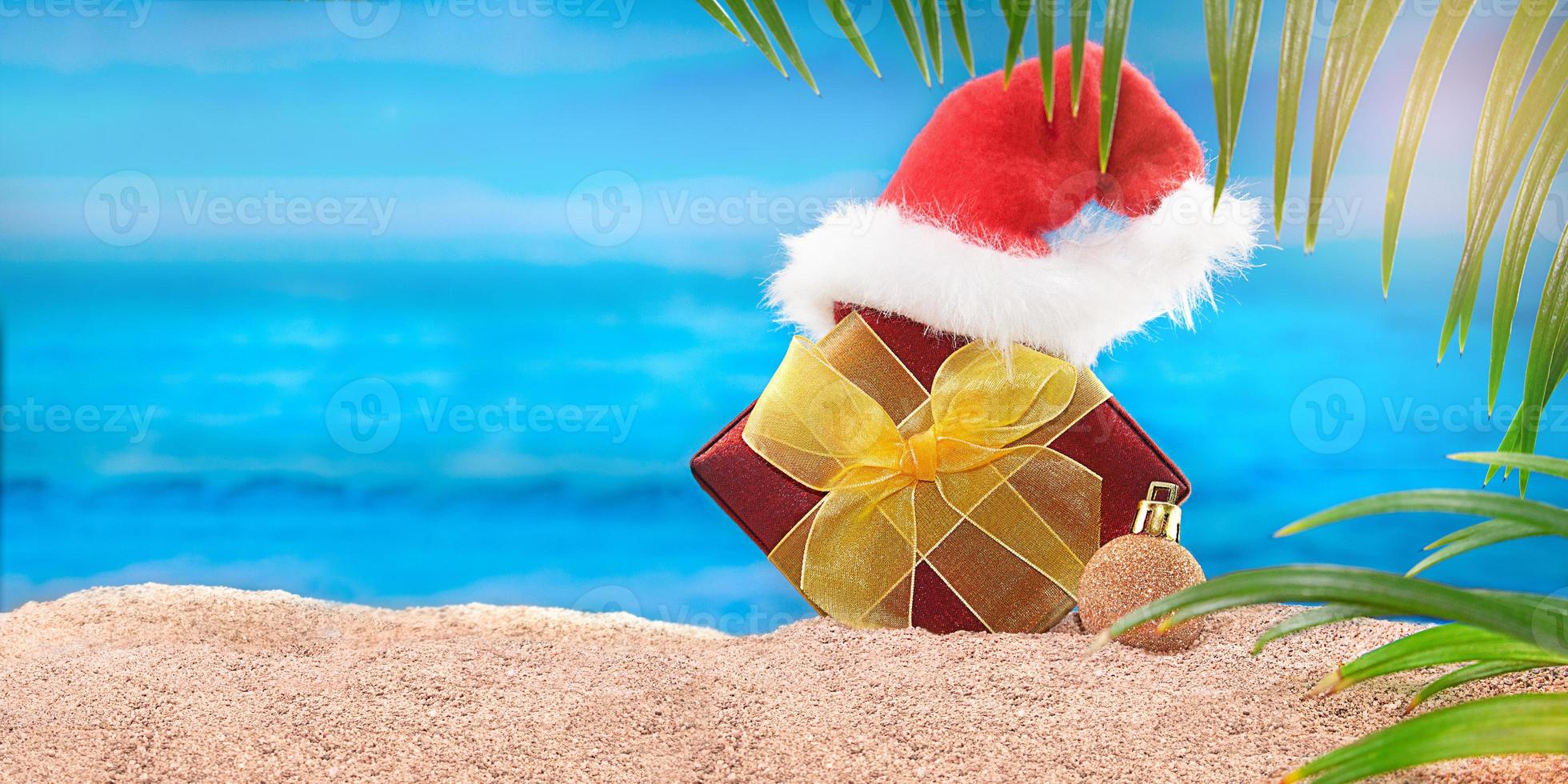 Red gift box with yellow bow and Santa Claus' hat on the beach behind sea and palm trees. Concept of Christmas, New year holiday in hot countries. Copy space photo