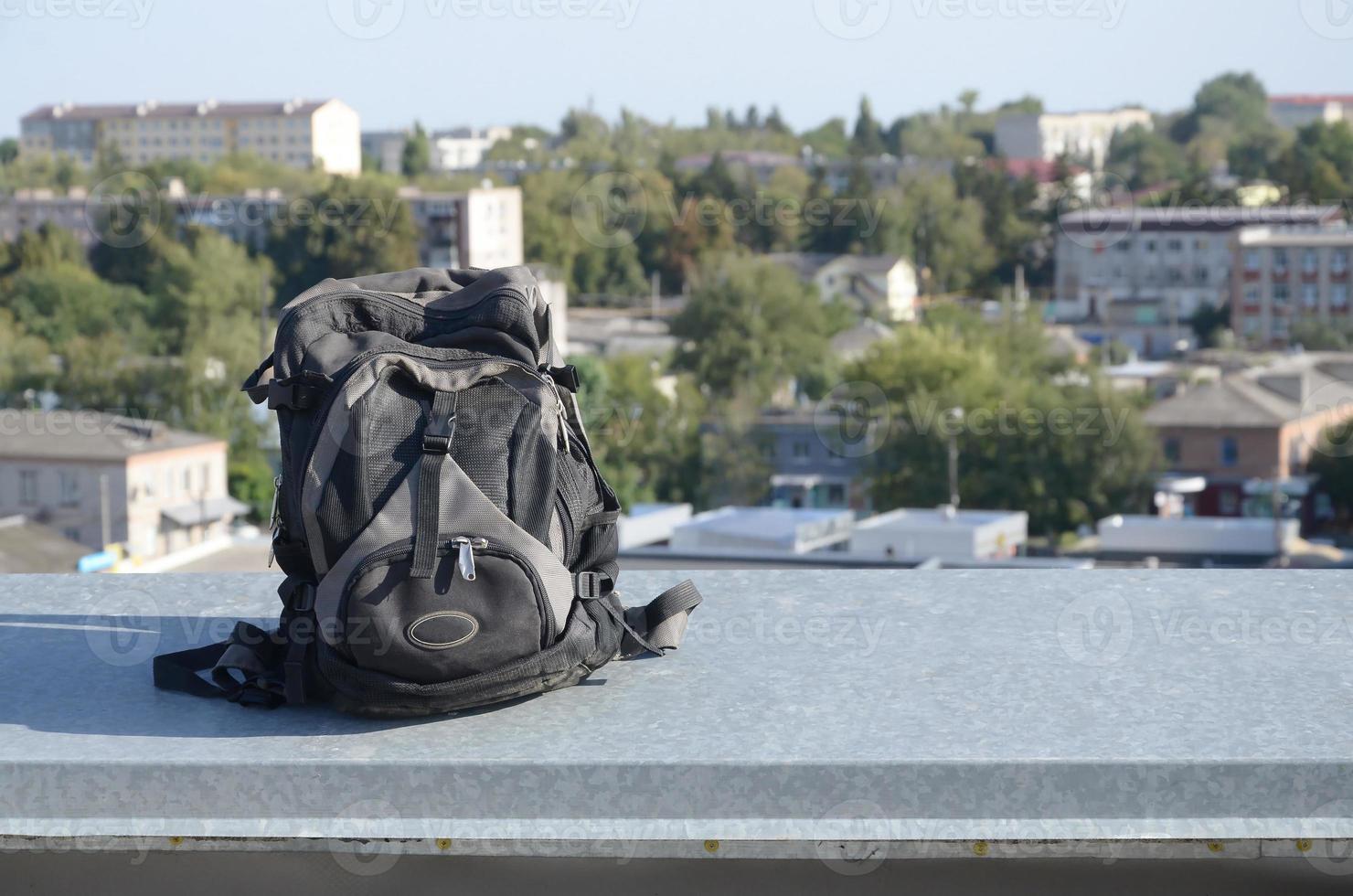 Black backpack lies on metal border of residental multistorey building rooftop in sunny weather photo