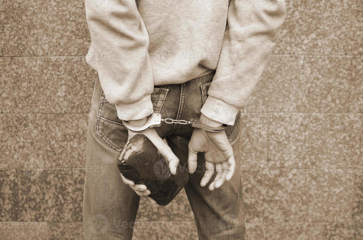 Arrested drug dealer in police handcuffs with big heroin drug package on dark wall background photo