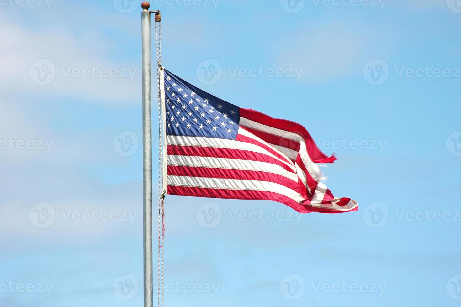 An old American flag on the tip of the flag with a torn in the wind on the flagpole against a blue sky background. photo