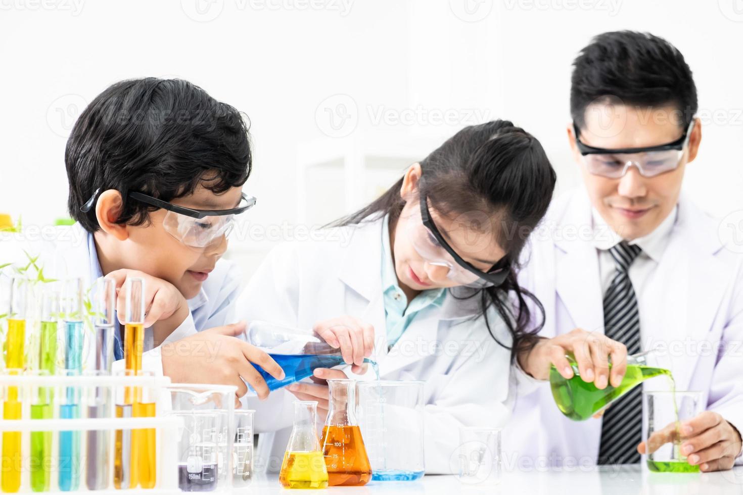 Young Asian kids wearing white scientist gown and do chemical test tube while study, learning in science classroom with teacher. Young children education concept by experiment, fun and enjoy classroom photo