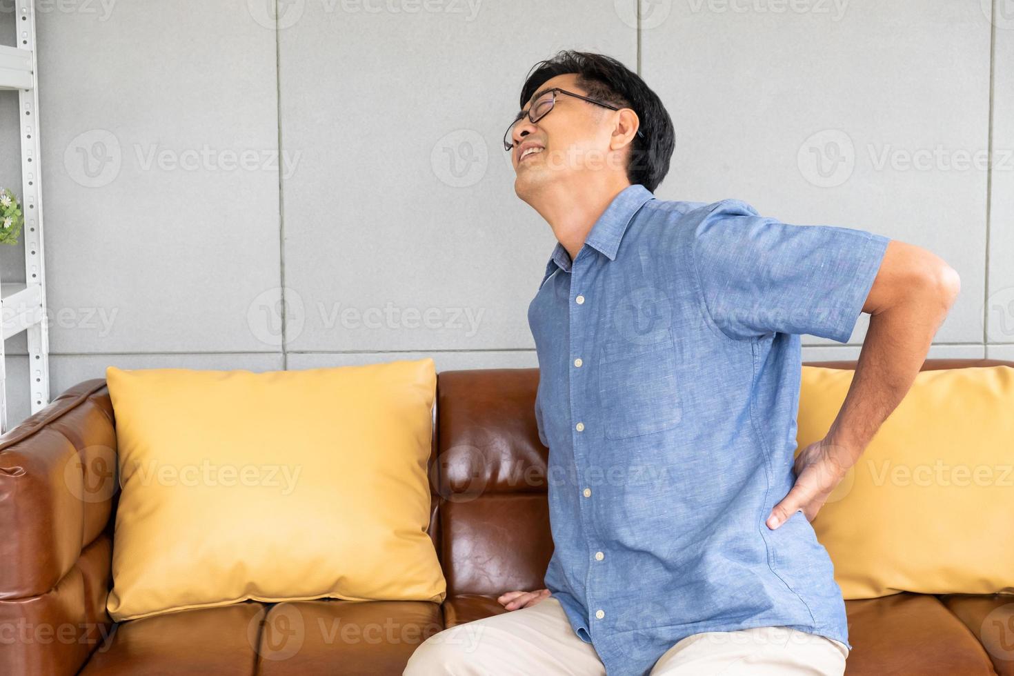 Indoor shot of senior Asian men sit on the sofa couch and touch his back and suffering from back pain, aching muscles, bones or lumbago. Elderly health problem concept. photo