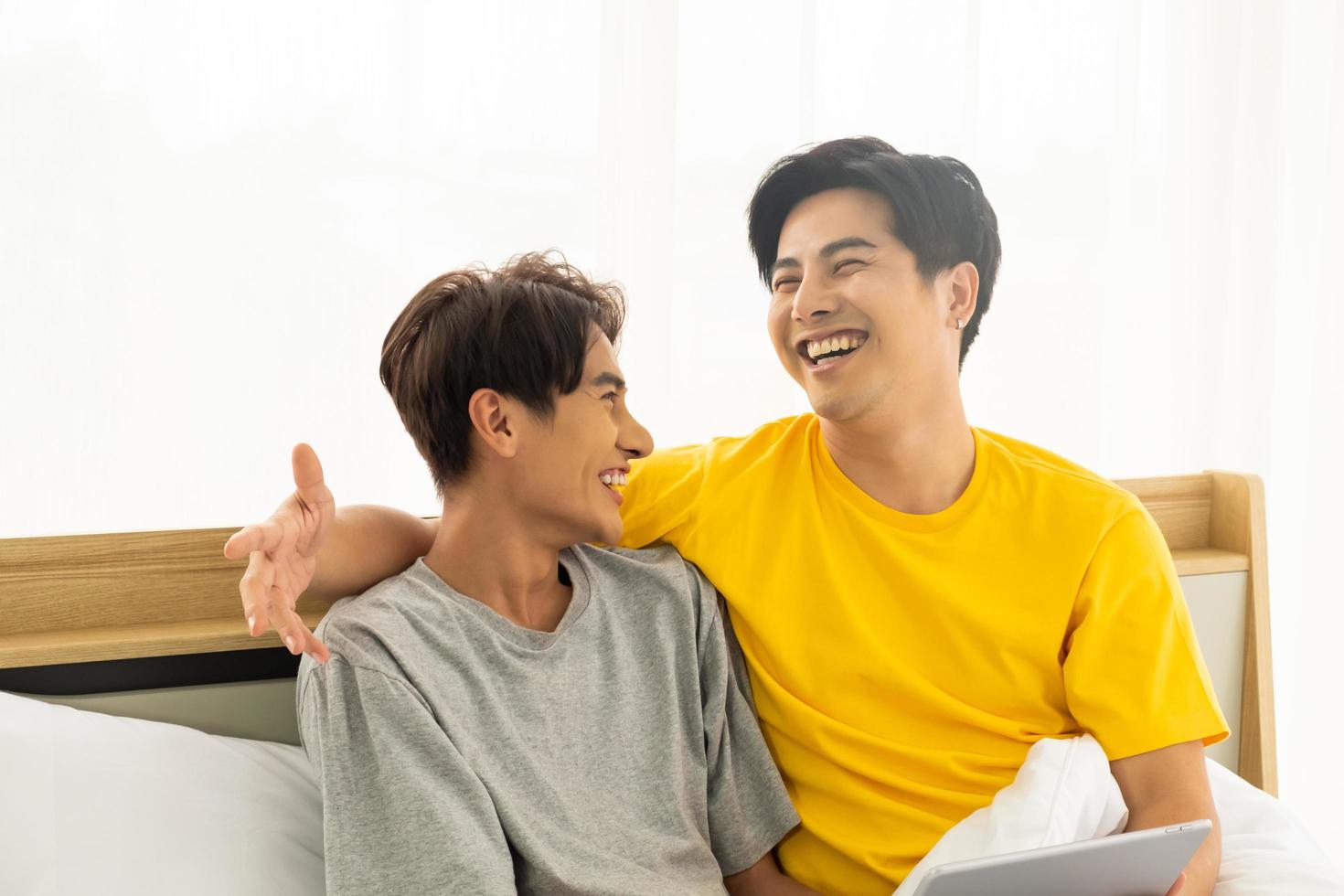 Selective focus at face. Good looking Asian gay homosexual couple hug and smile while using Digital tablet smartphone, showing love expression on the bed. LGBT relationship concept with copy space. photo