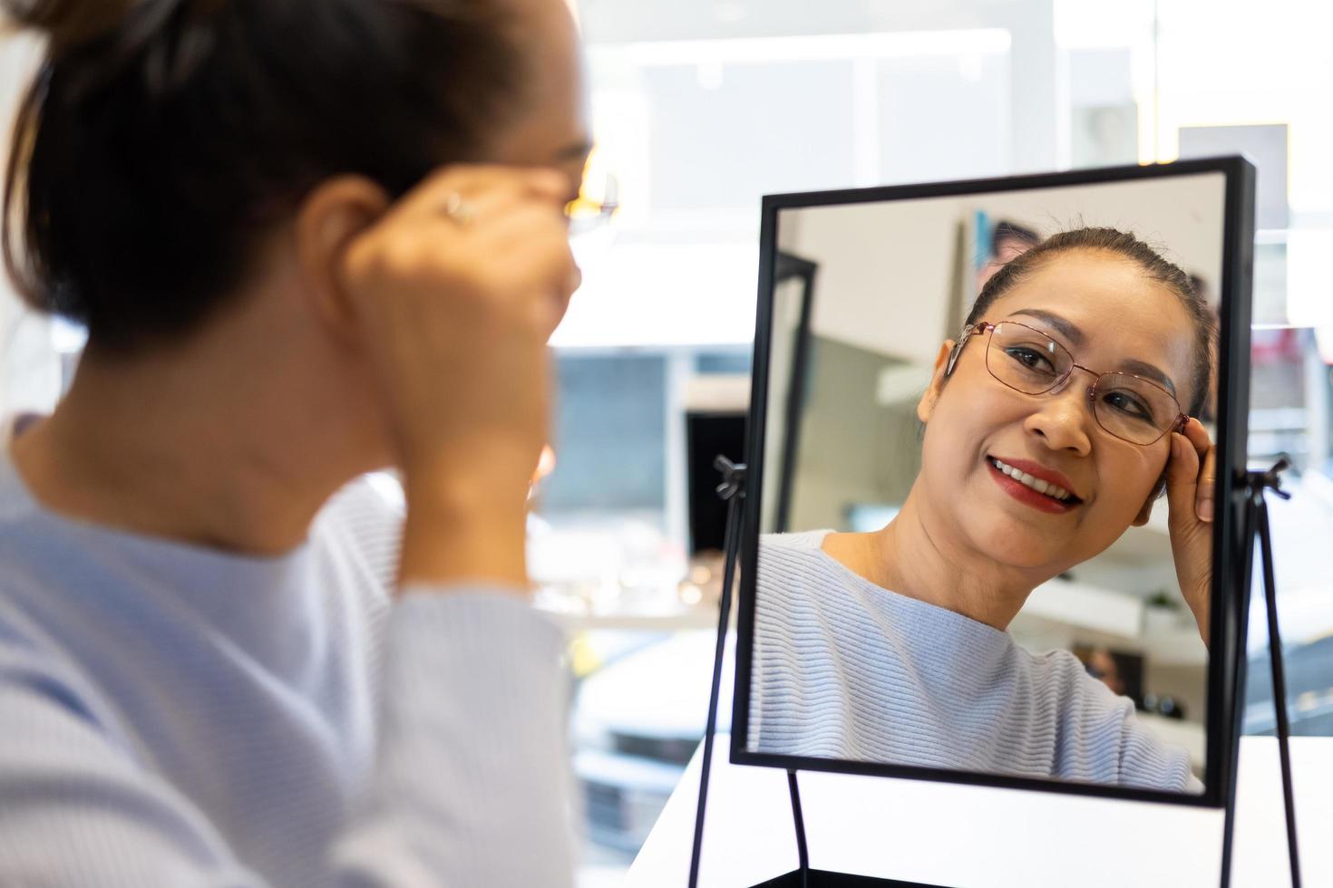 Selective focus at female face. Senior elder Asian women smile and look at the mirror while choosing beautiful glasses frame trial inside of optical shop, store.  Eye sight for elder, healthcare. photo