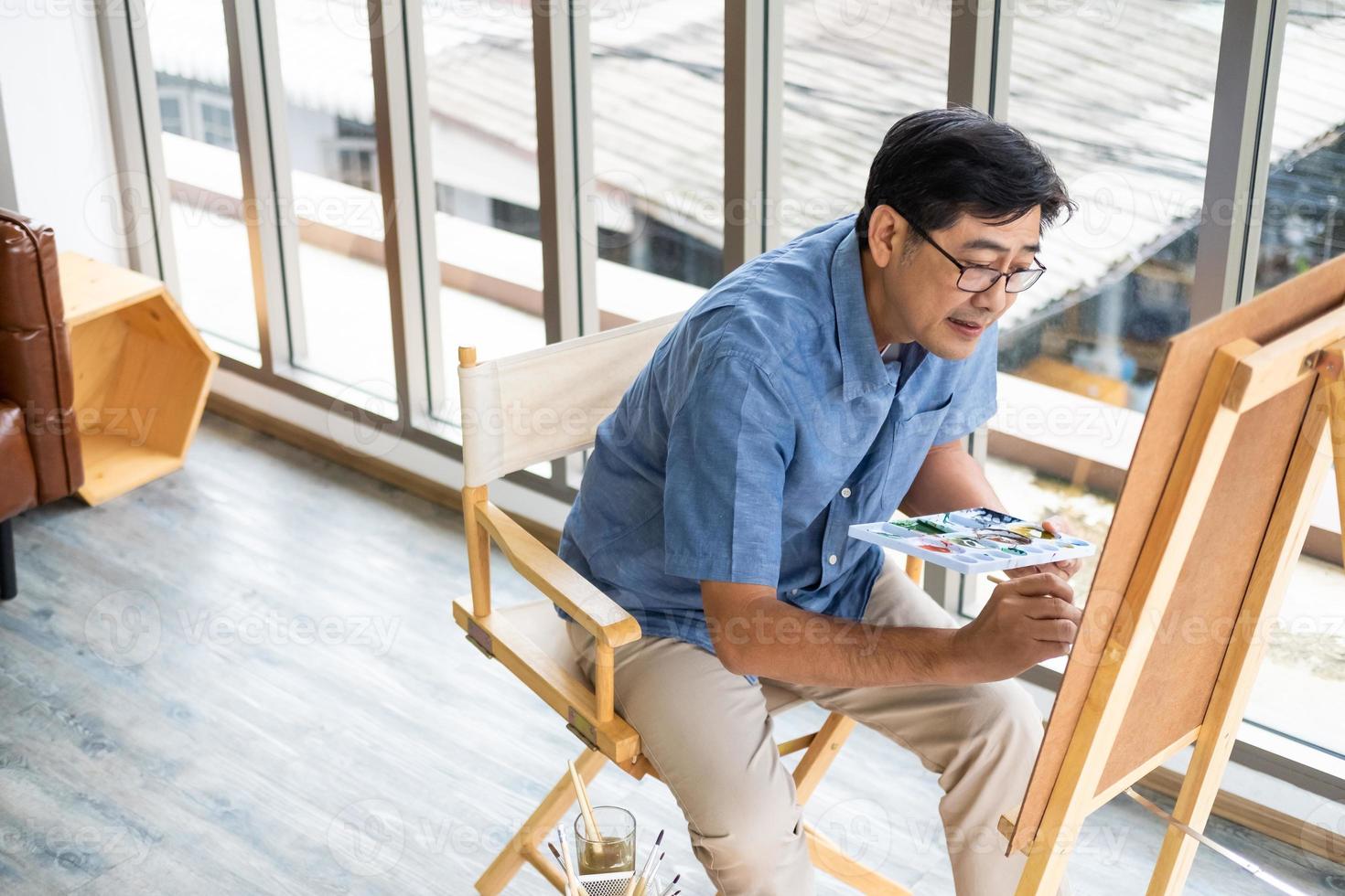 Selective focus at face. Indoor shot of Senior Asian man sit and painting picture inside of the house. Happy retirement lifestyle, active elderly with copy space. photo