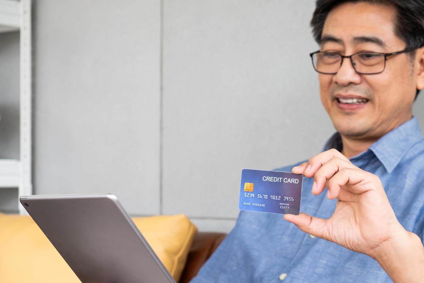 Selective focus at credit card. Indoor shot of senior Asian men sit down on sofa and using computer laptop for online credit card payment with smile on his face. Happy senior with technology. photo