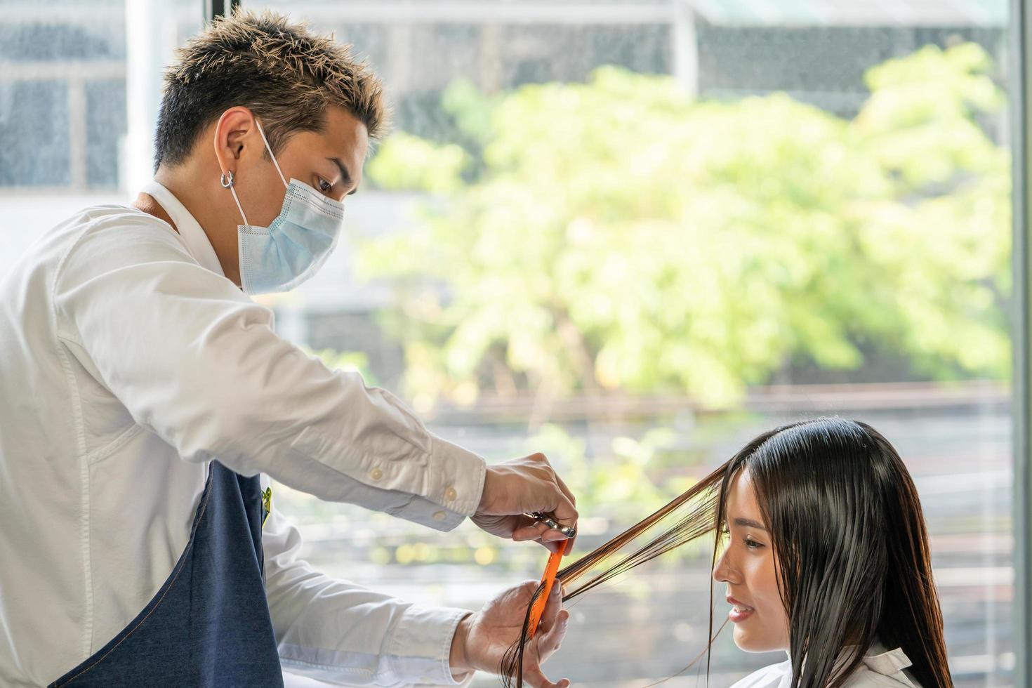 Selective focus at face of Asian women with Hair stylish while do hair cut and wearing surgical face mask while styling hair for client. Professional occupation, beauty and fashion service new normal photo