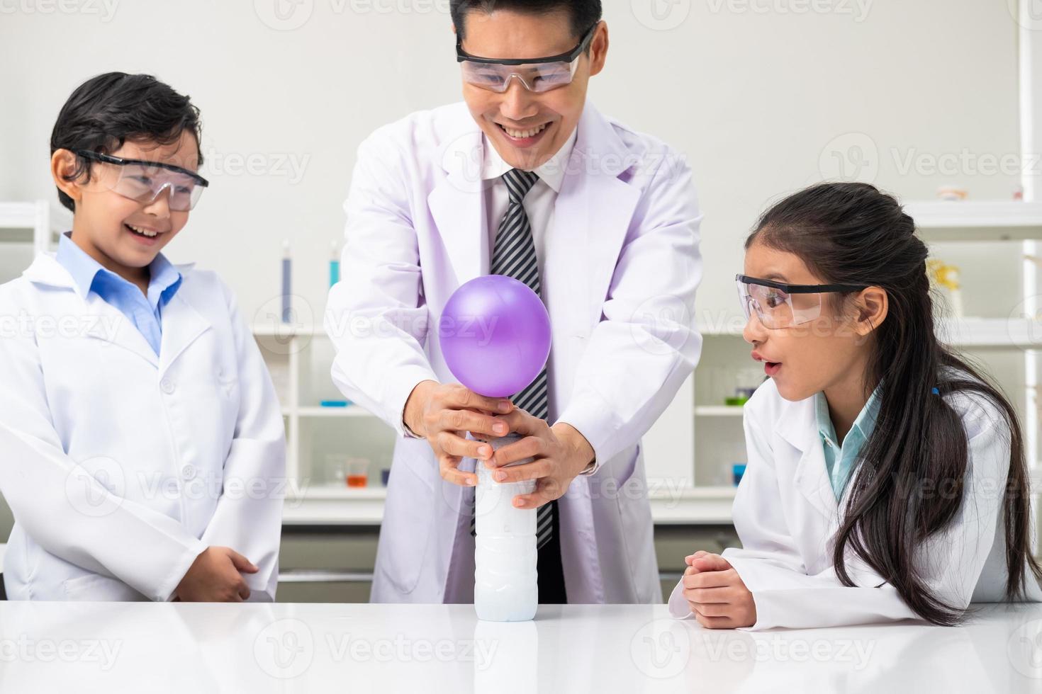 Young Asian kids wearing white scientist gown and do chemical test tube while study, learning in science classroom with teacher. Young children education concept by experiment, fun and enjoy classroom photo