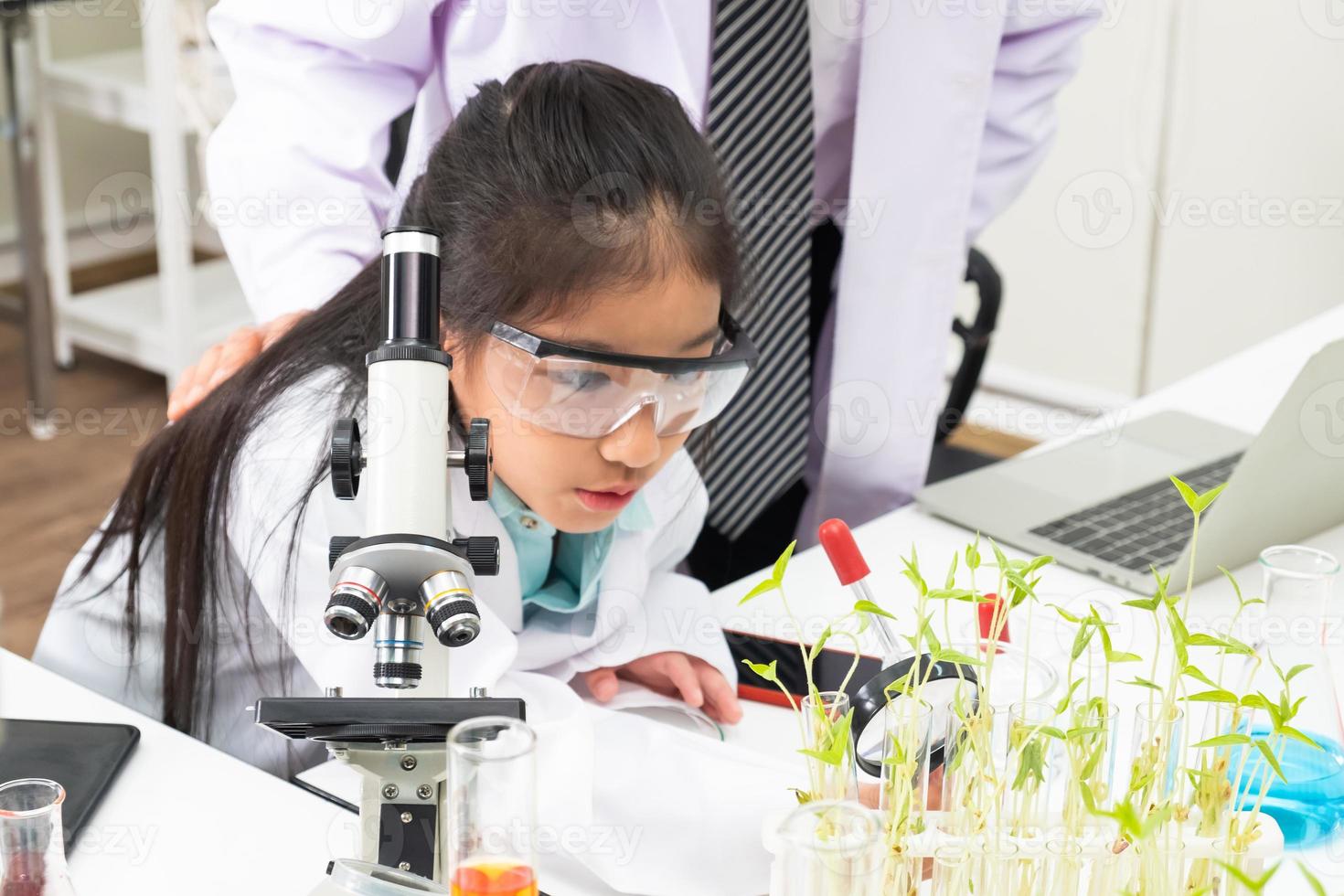 un joven estudiante de elementos asiáticos se ajusta y usa un microscopio en la clase de ciencias para examinar el tema de la vida con el maestro. educación y niños que aprenden o experimentan en el concepto de aula. foto