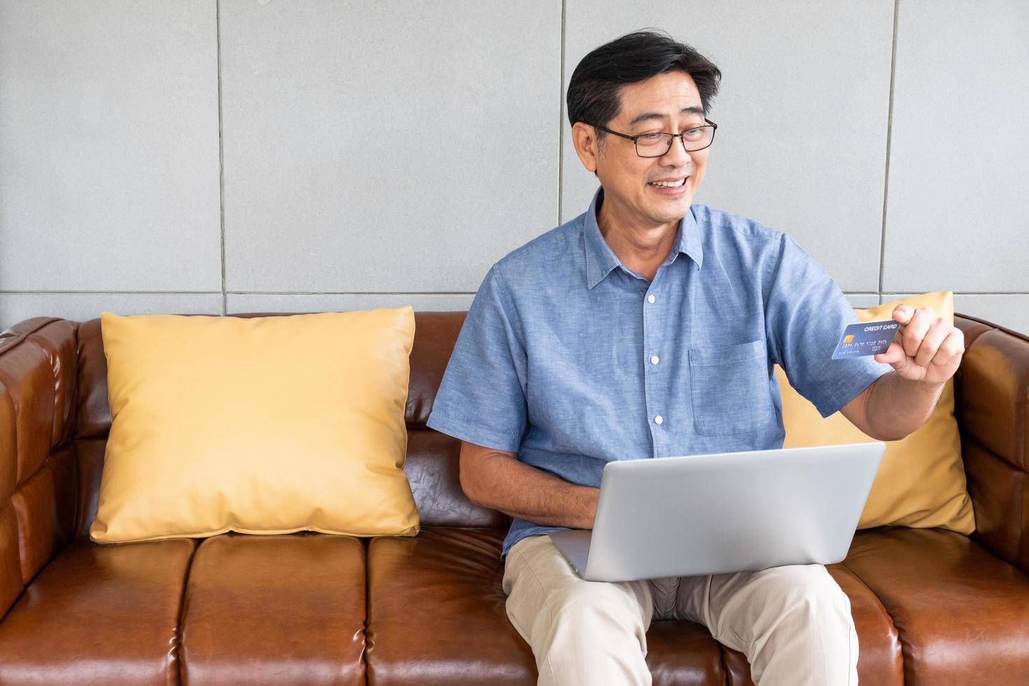 Indoor shot inside of the house of senior Asian men sit down on sofa and using computer laptop for online credit card payment with smile on his face. Happy senior with technology. photo