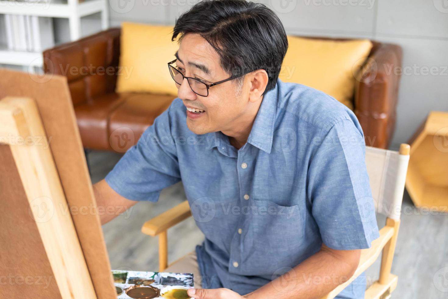 Indoor shot of senior Asian men using brush and oil color to paint on painting canvas. With smile on his face. Happy retirement concept for active senior. With Blurred living room background. photo
