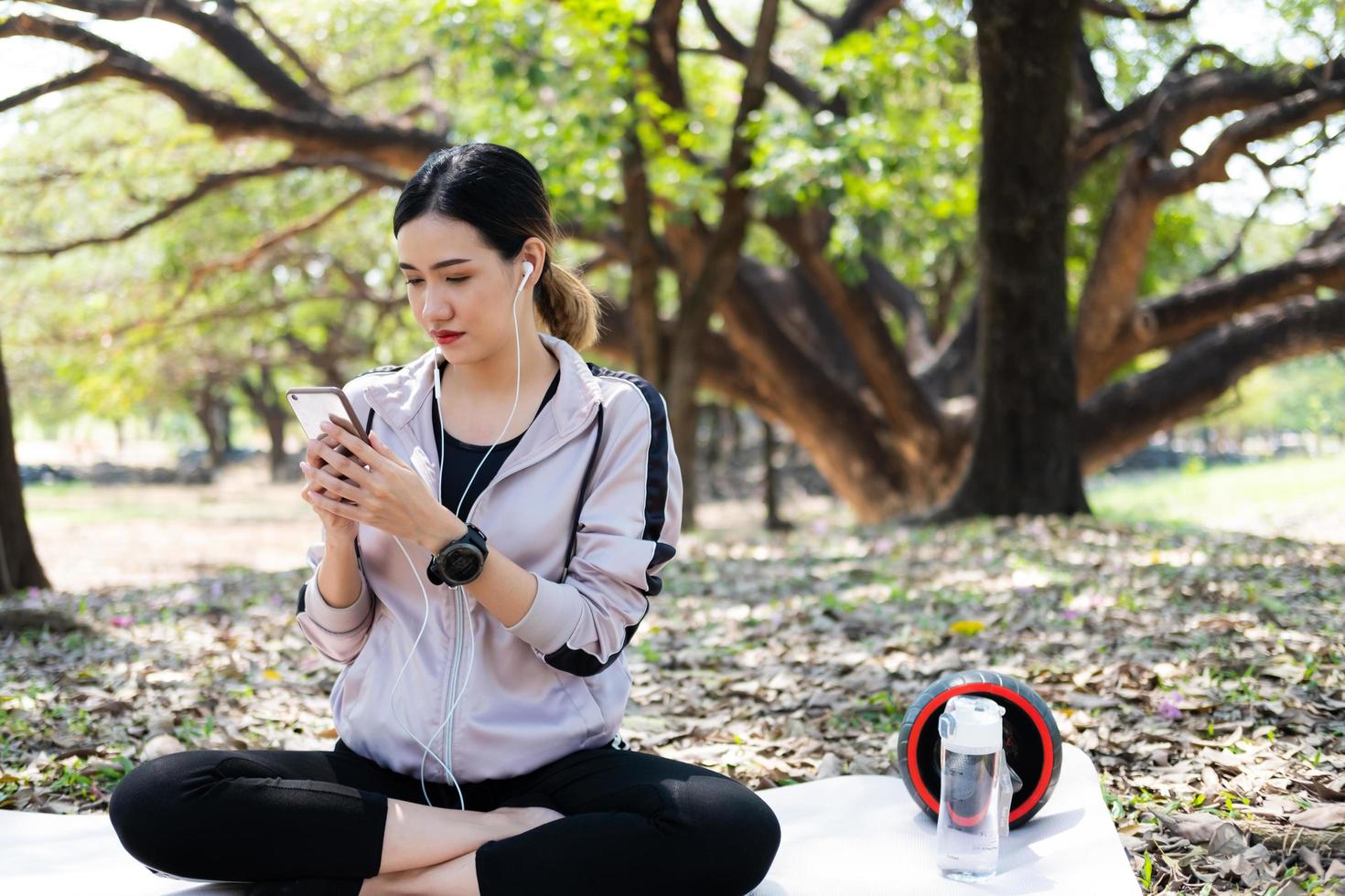 enfoque selectivo en la cara de las jóvenes y hermosas mujeres asiáticas que usan un reloj inteligente para rastrear la actividad y escuchar música desde el teléfono inteligente mientras se calientan antes de hacer ejercicio de yoga con árboles naturales y de fondo. foto