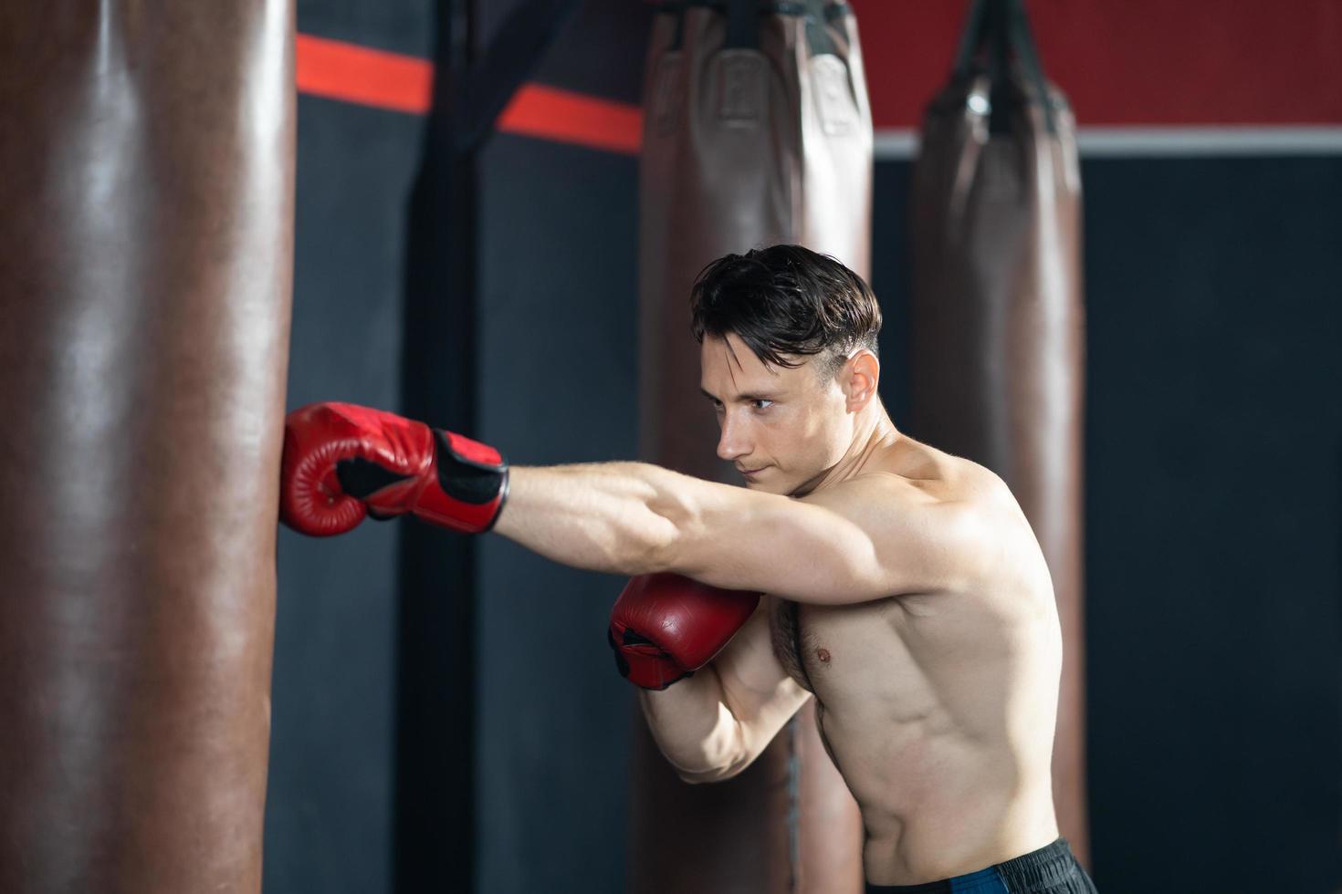 Shirtless handsome Caucasian men boxer with strong muscle training boxing punch in side of the gym with sandbag. Cardio sport workout activity for good health and build up body and physical strength. photo