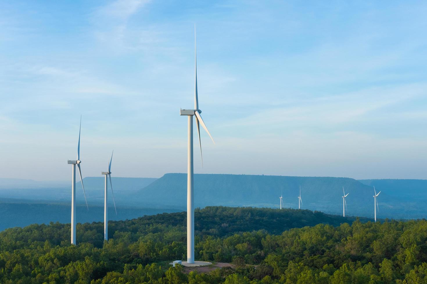 Landscape sun set shot of Wind turbines farm on the green grass field with clear blue sky and mountain at the background. Alternative green energy which generate electricity from natural wind power. photo