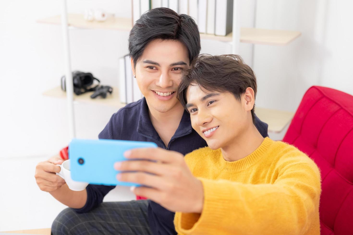 Young Asian handsome and good looking homosexual couple using smartphone to taking selfie photo together while sitting on sofa inside living room. Male, same sex relationship lifestyle.