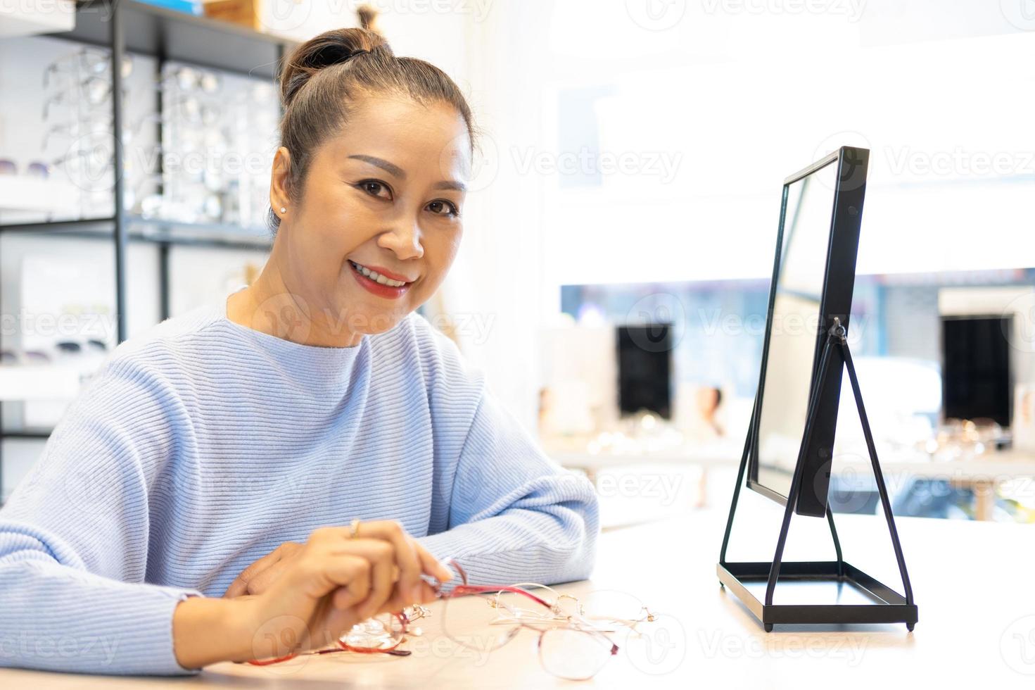 Selective focus at the face.  Senior elder Asian women smile and look at the mirror while choosing beautiful glasses frame trial inside of optical shop, store.  Eye sight for elder, healthcare. photo