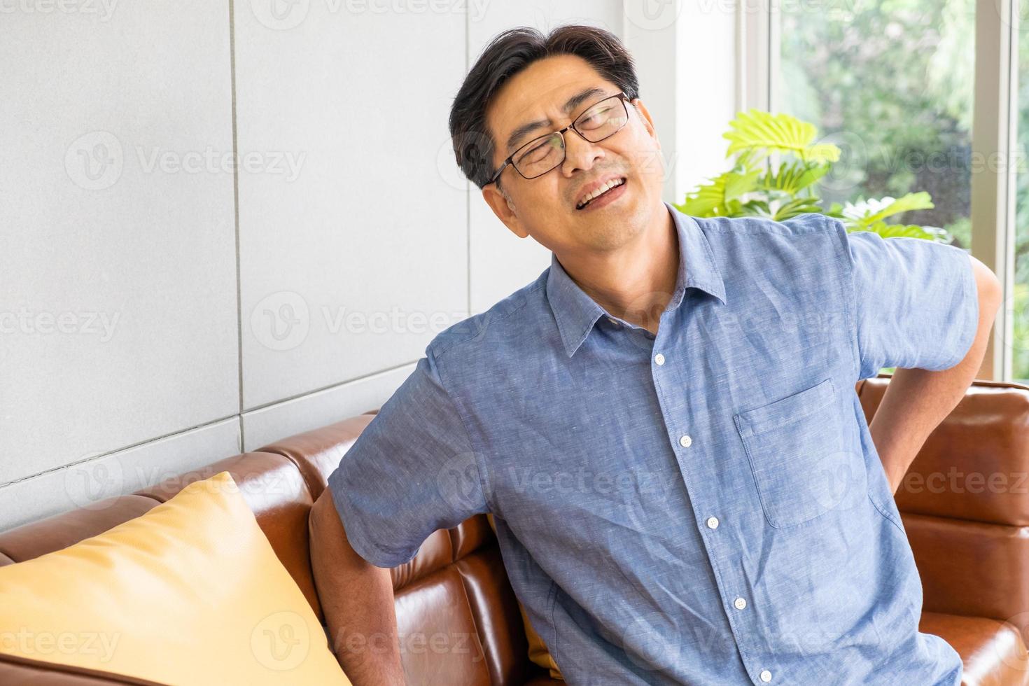 Indoor shot of senior Asian men sit on the sofa couch and touch his back and suffering from back pain, aching muscles, bones or lumbago. Elderly health problem concept. photo