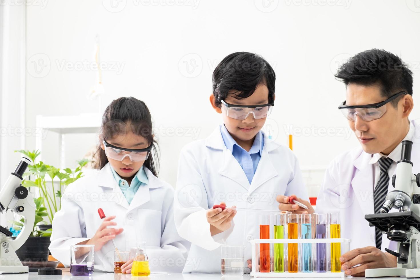 Young Asian kids wearing white scientist gown and do chemical test tube while study, learning in science classroom with teacher. Young children education concept by experiment, fun and enjoy classroom photo