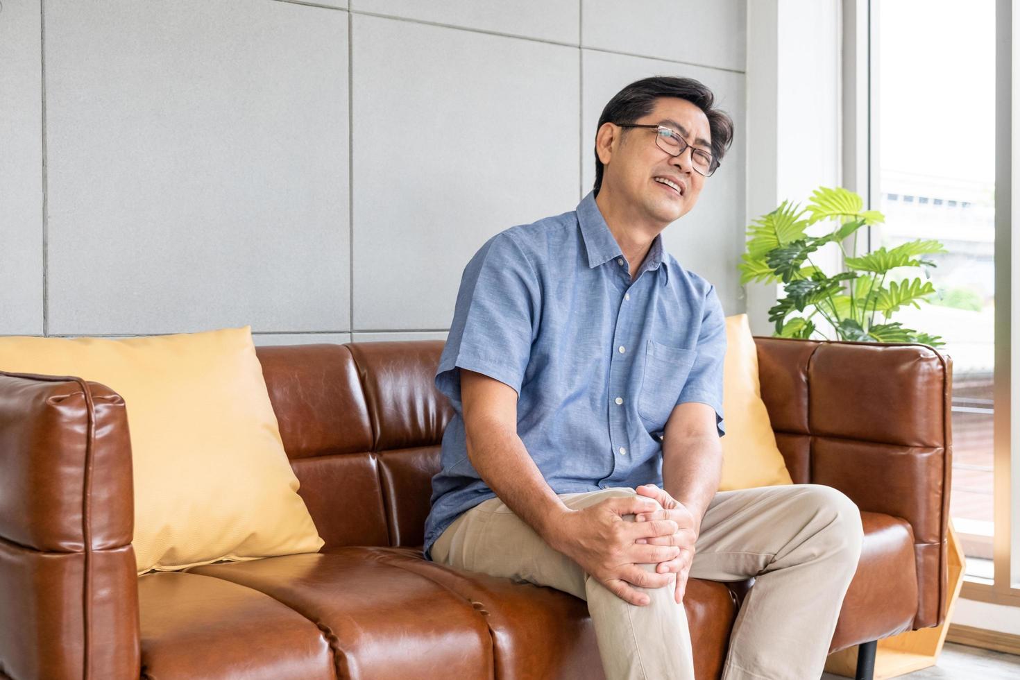Selective focus at face of old senior Asian man sitting at sofa touching knee and  legs suffer from the pain and injury. Healthcare and medical problem of Elder people concept. photo