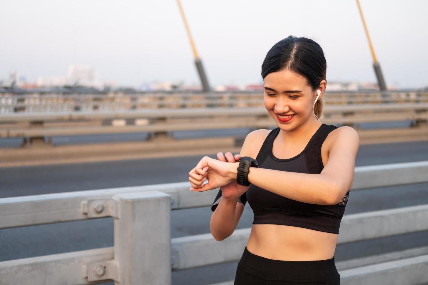 Selective focus at face of young beautiful Asian women using smart watch to track activity and listen music while warm up before exercise or running at the urban city view. Fit and healthy lifestyle. photo