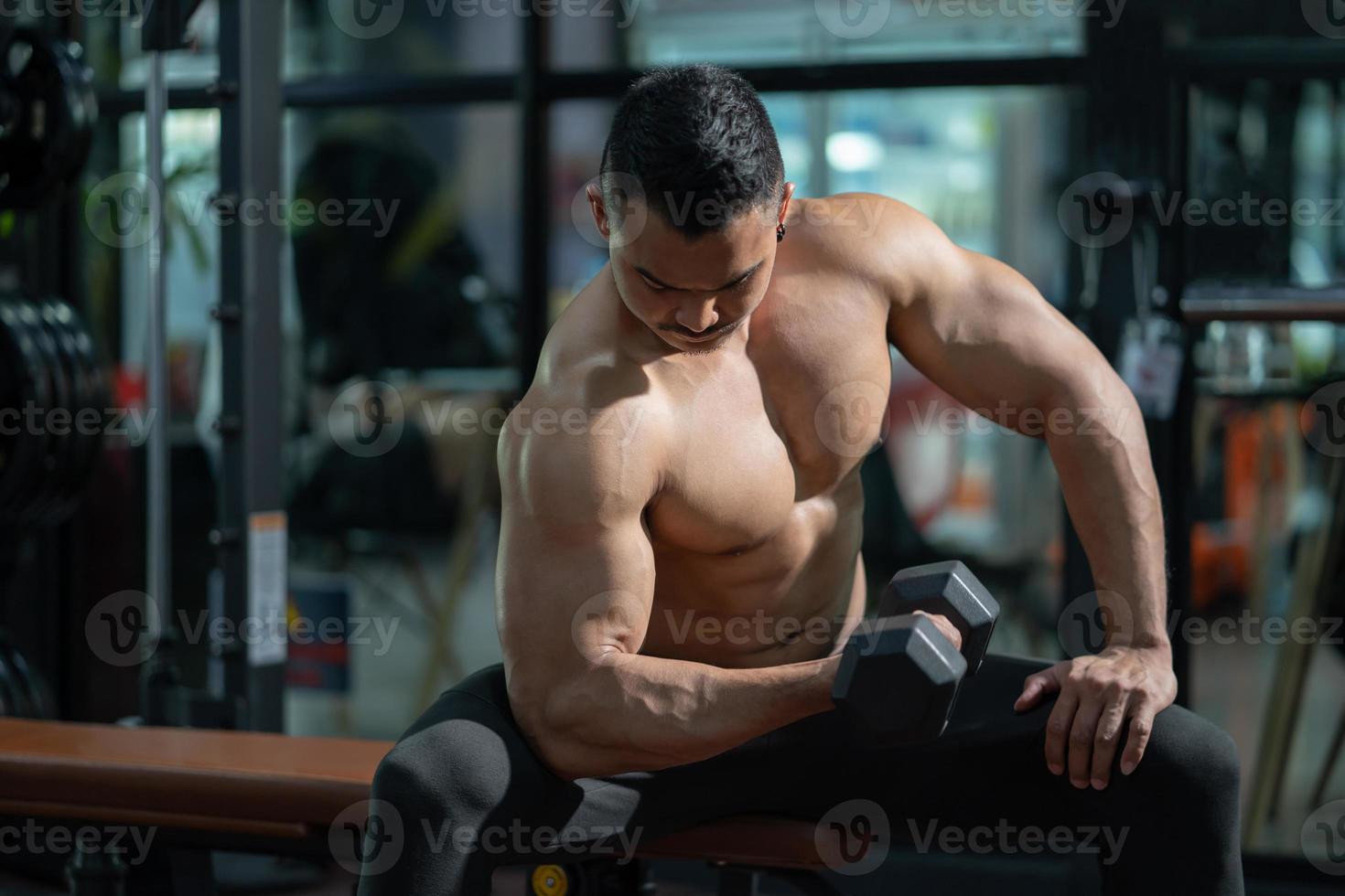 Handsome shirtless adult Asian men sweating while lift up the dumbbell workout for arm biceps muscle part inside of fitness gym. Bodybuilding athlete sport training for body strength and good health. photo