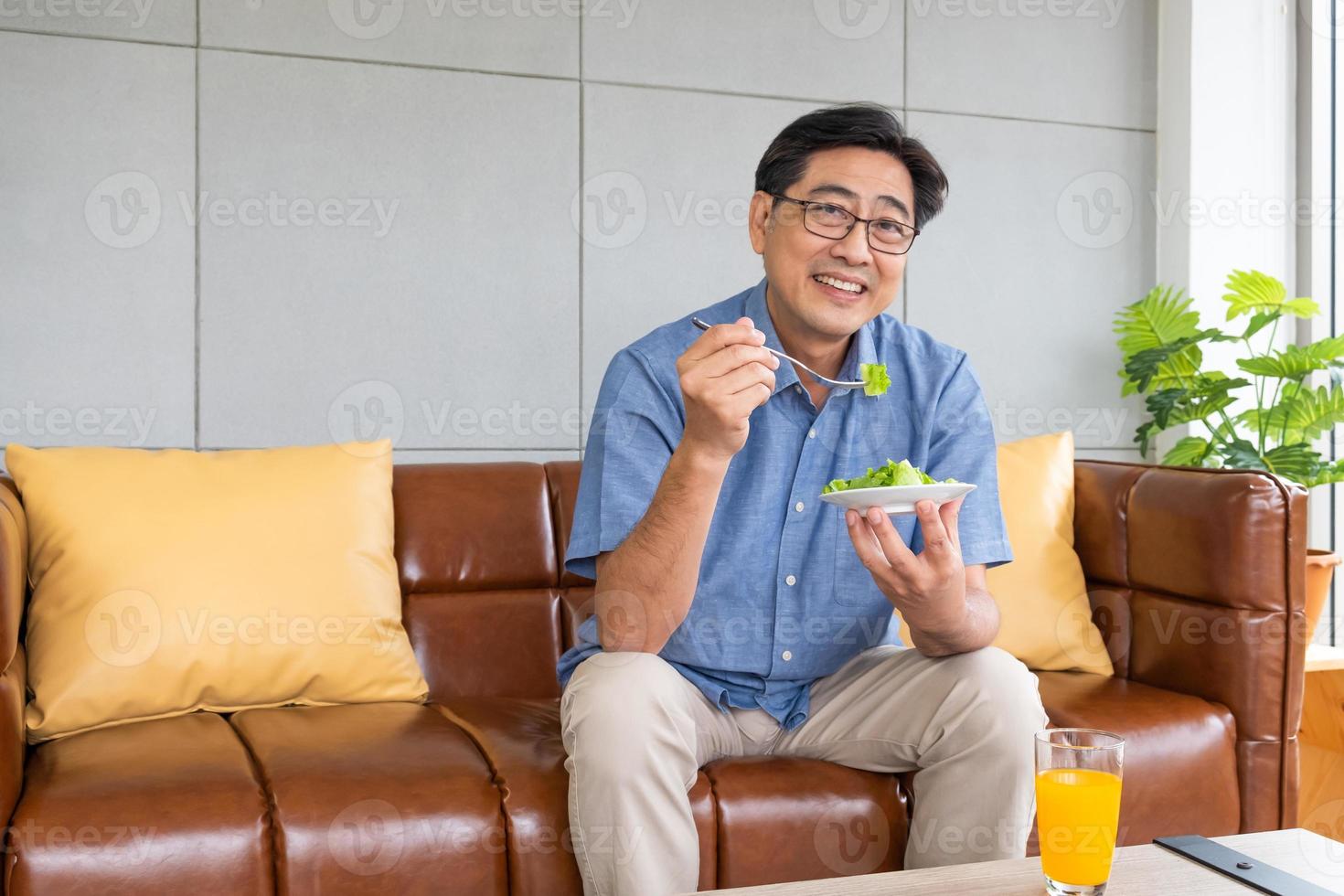 Senior Asian men sitting on the sofa at home while eating green vegetable salad and organic orange juice for breakfast. Healthy eating lifestyle for Elder people concept. photo