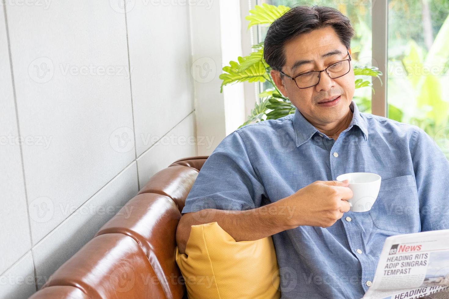 Selective focus at face. Senior Asian man reading newspaper mock up at the sofa couch while drinking coffee in the morning. Happy retirement with relaxation lifestyle and routine. photo