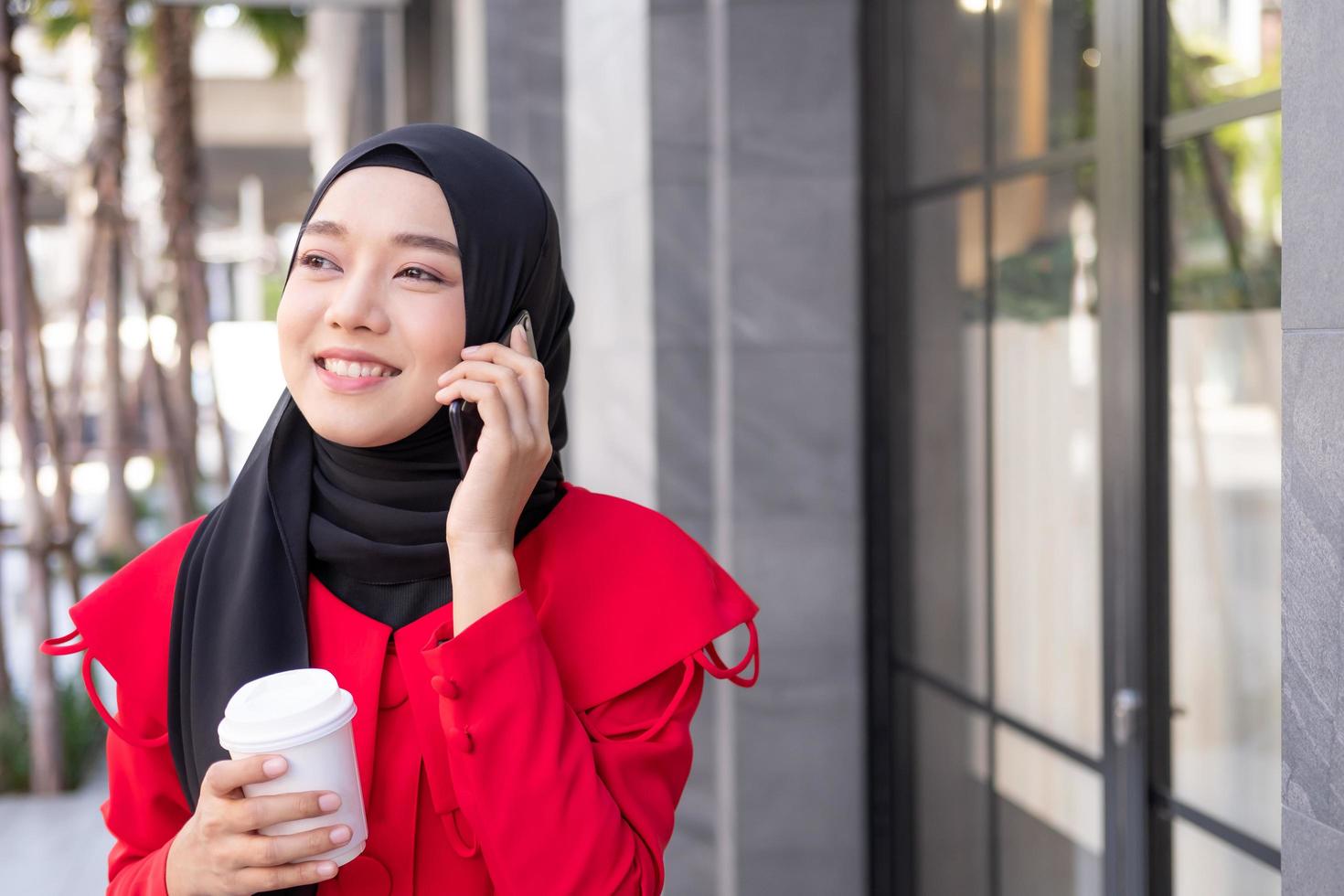 hermosas mujeres islámicas del este de Asia usando hiyab con vestido formal. hablando por teléfono móvil mientras sostiene una taza de café y camina en el área urbana de la ciudad o en los grandes almacenes. concepto de estilo de vida de las personas. foto