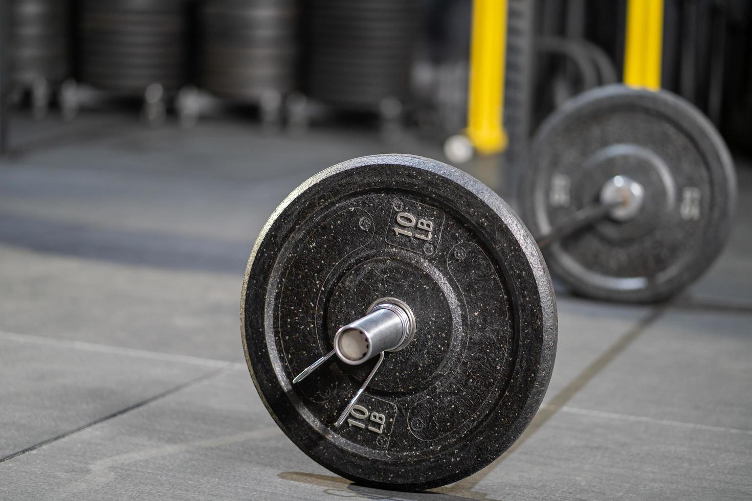Selective focus at the iron weight plate attached to barbell on the floor inside of fitness gym with blurred background of weight lifting machine for muscle build with copy space. Sport object concept photo