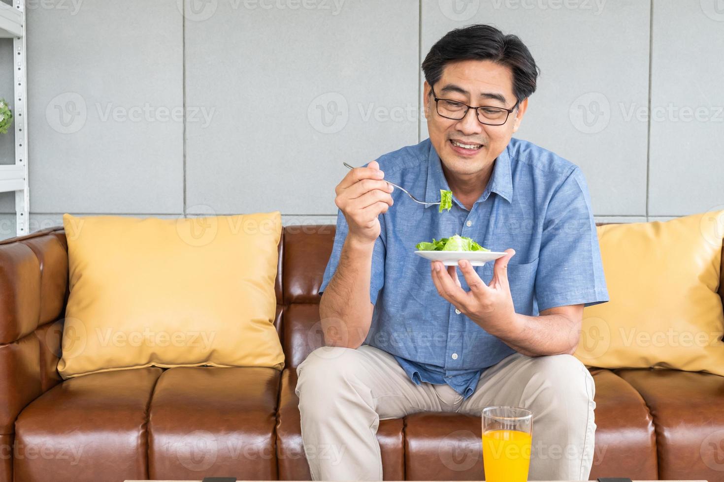 Senior Asian men sitting on the sofa at home while eating green vegetable salad and organic orange juice for breakfast. Healthy eating lifestyle for Elder people concept. photo